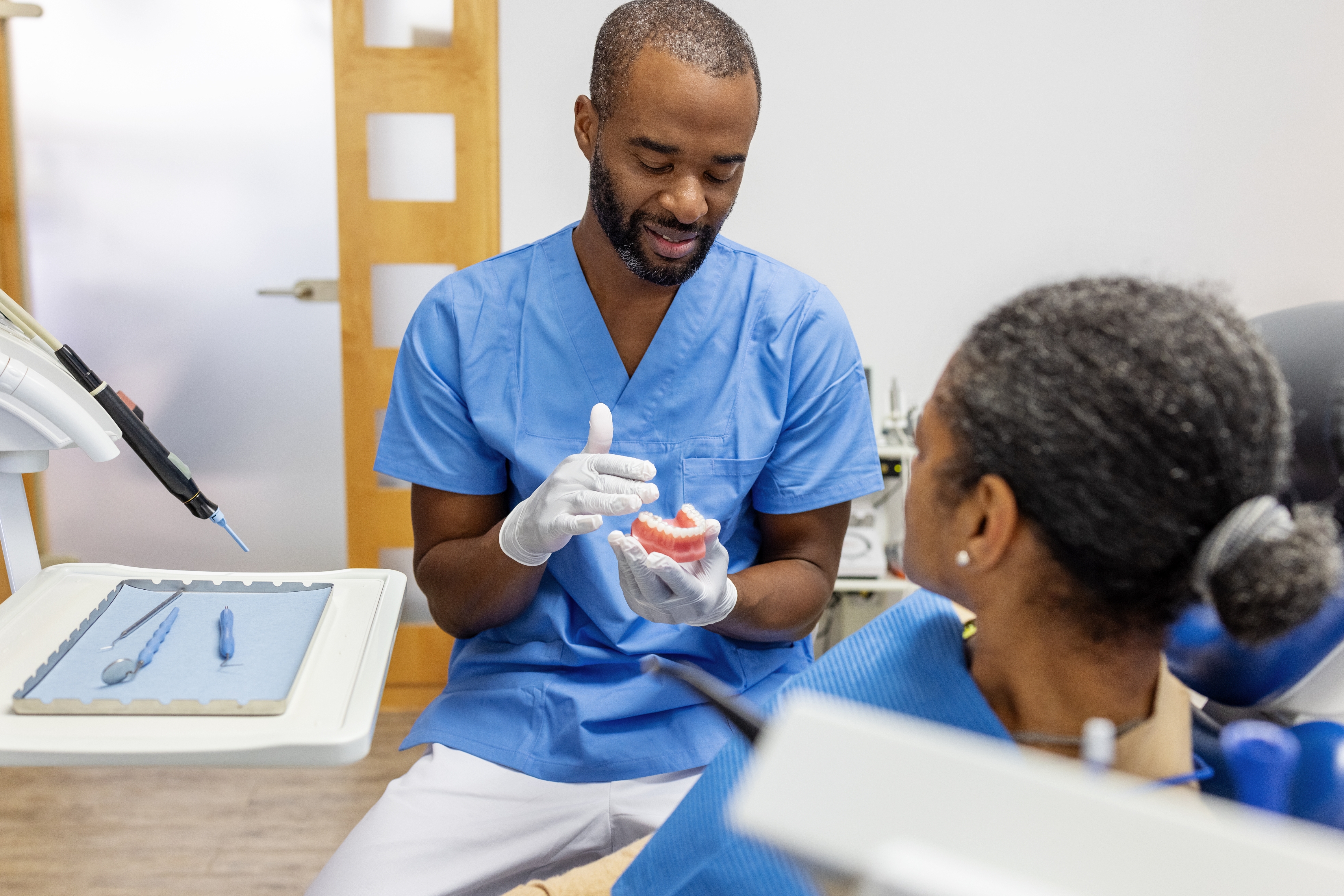 Dentist in scrubs shows a dental model to a seated patient, explaining a procedure in an exam room