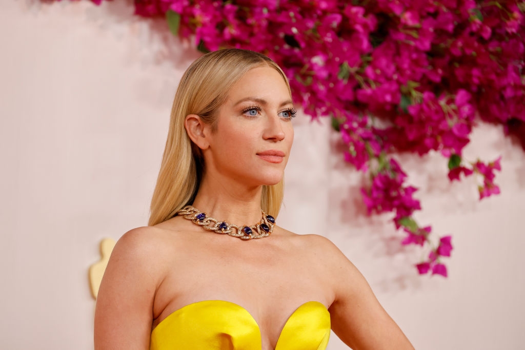 Celebrity on red carpet in elegant strapless gown with bold statement necklace; floral background enhances the scene