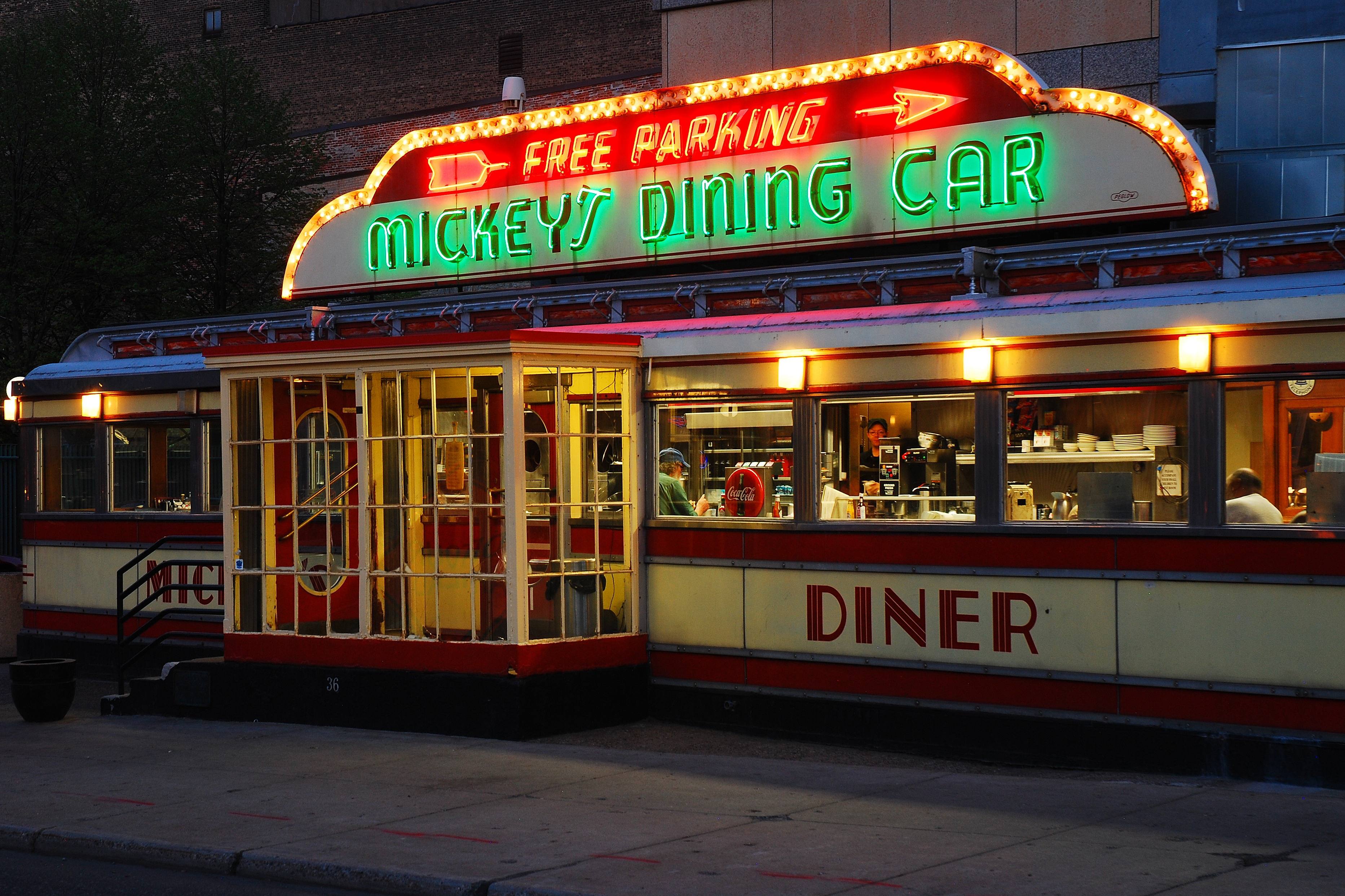 Retro diner with neon "Mickey's Dining Car" sign, warm interior lighting, and patrons visible inside