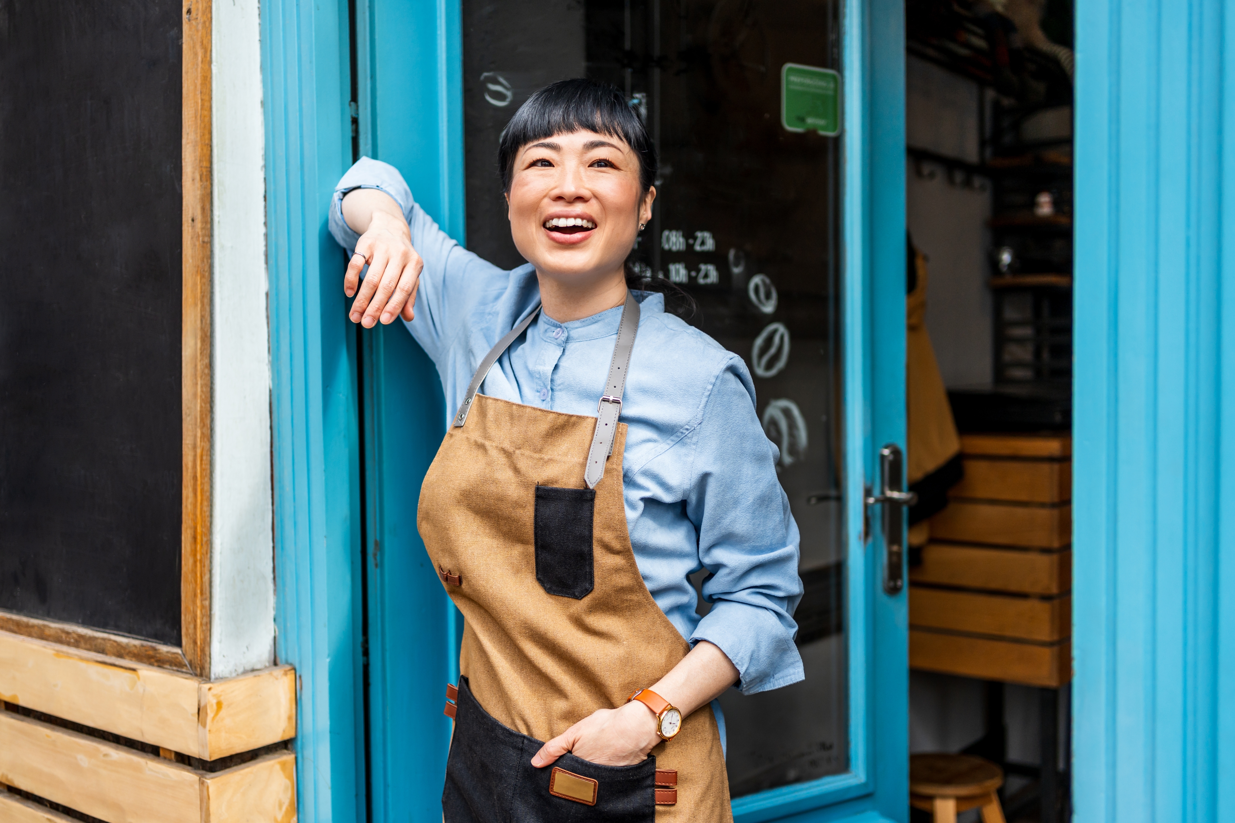 Person smiling, leaning on a bakery door, wearing a brown apron over a blue shirt