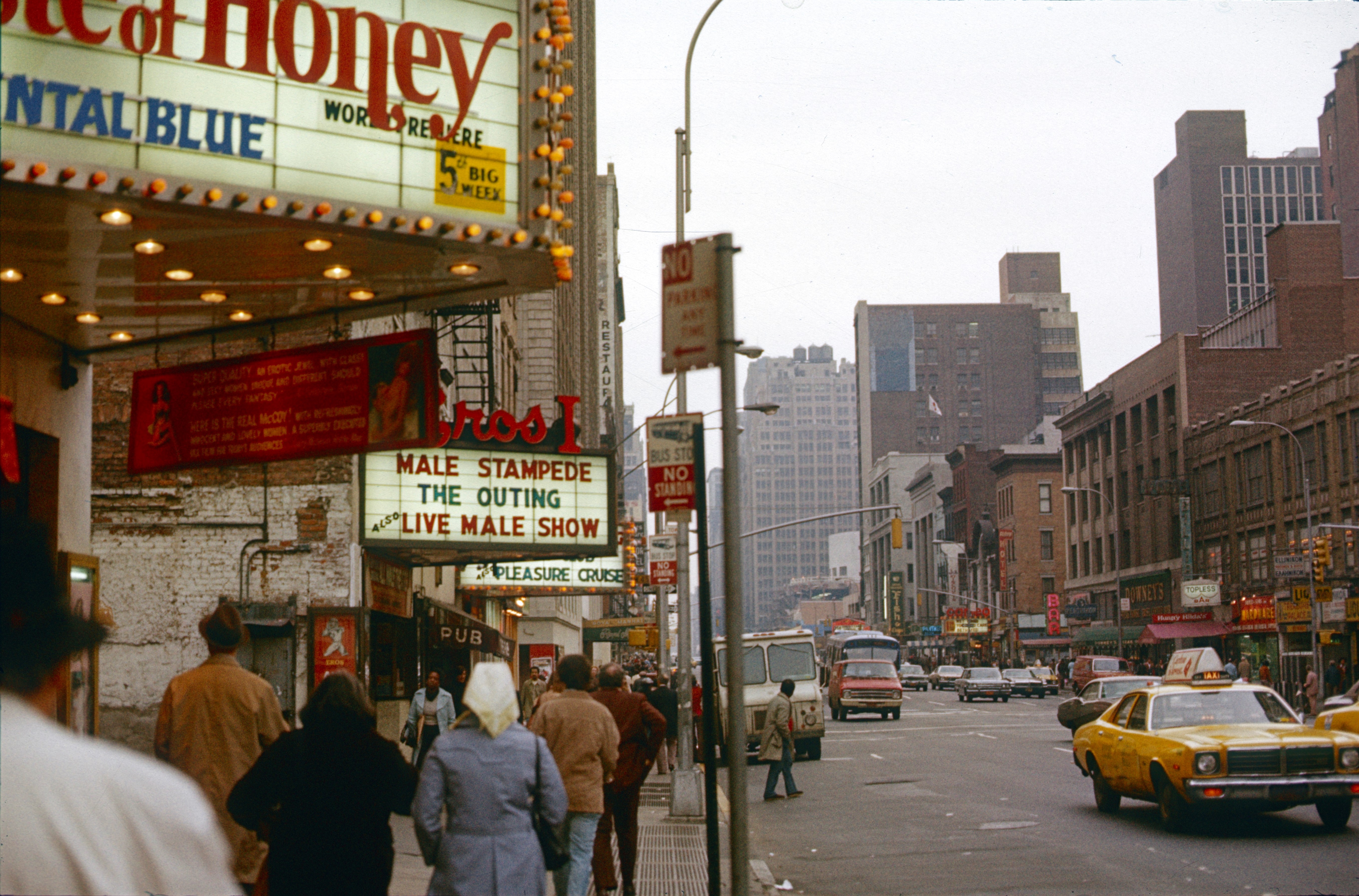 People walk down a busy city street with theater marquees advertising various shows, and cars drive by, including a yellow taxi