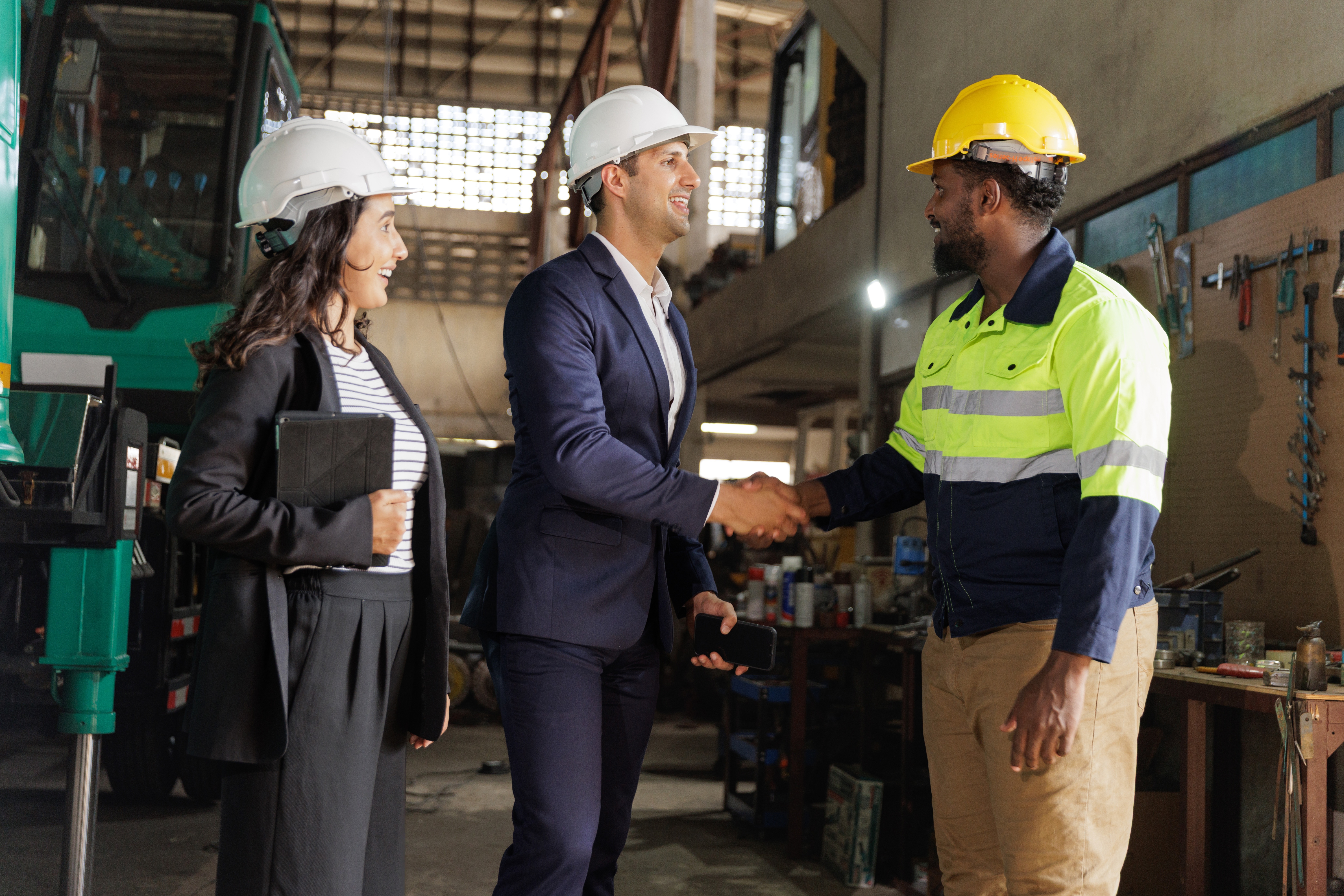 Three people wearing hard hats in a factory. Two in business attire shake hands with one in safety gear. They appear to be having a positive meeting