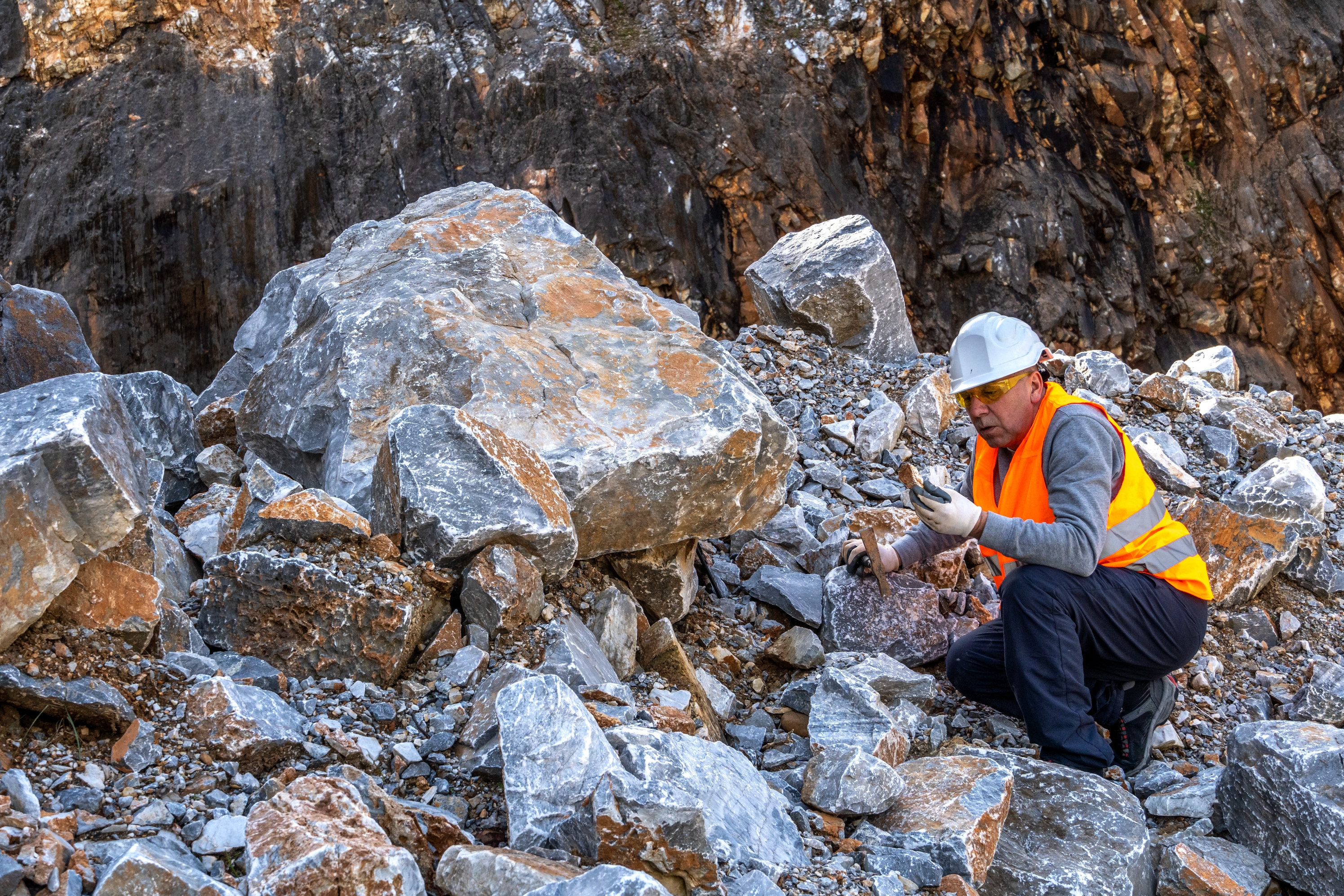 Worker in safety gear examines rocks in a quarry setting, surrounded by large boulders