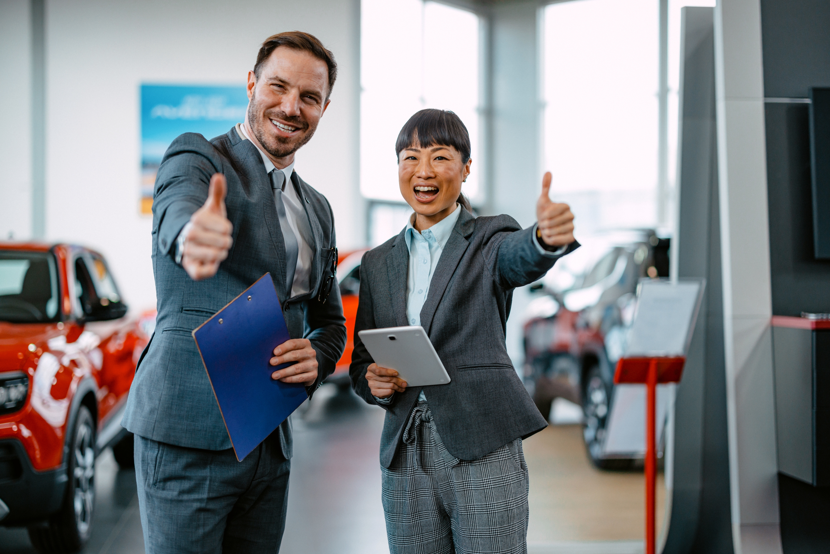 Two people in business attire smiling and giving thumbs up inside a car showroom, surrounded by vehicles