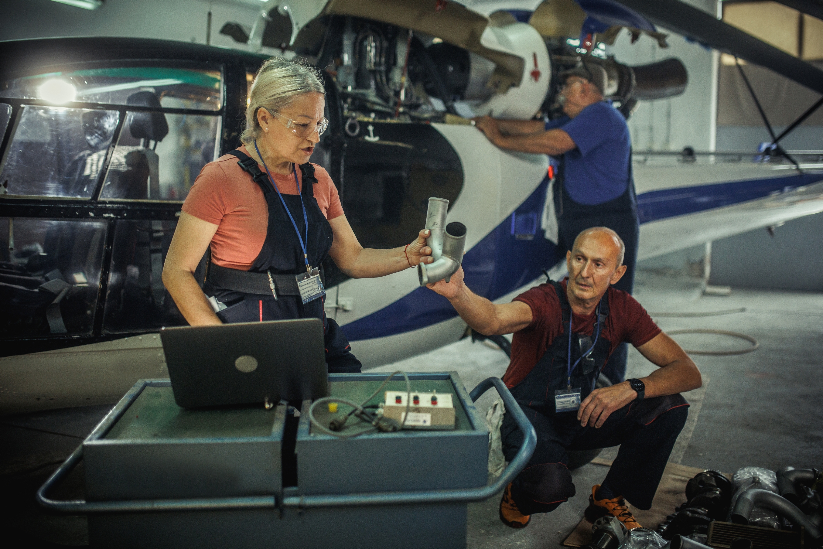 Two mechanics, one standing and one crouching, work together on an airplane's engine in a hangar, using tools and a laptop for repairs
