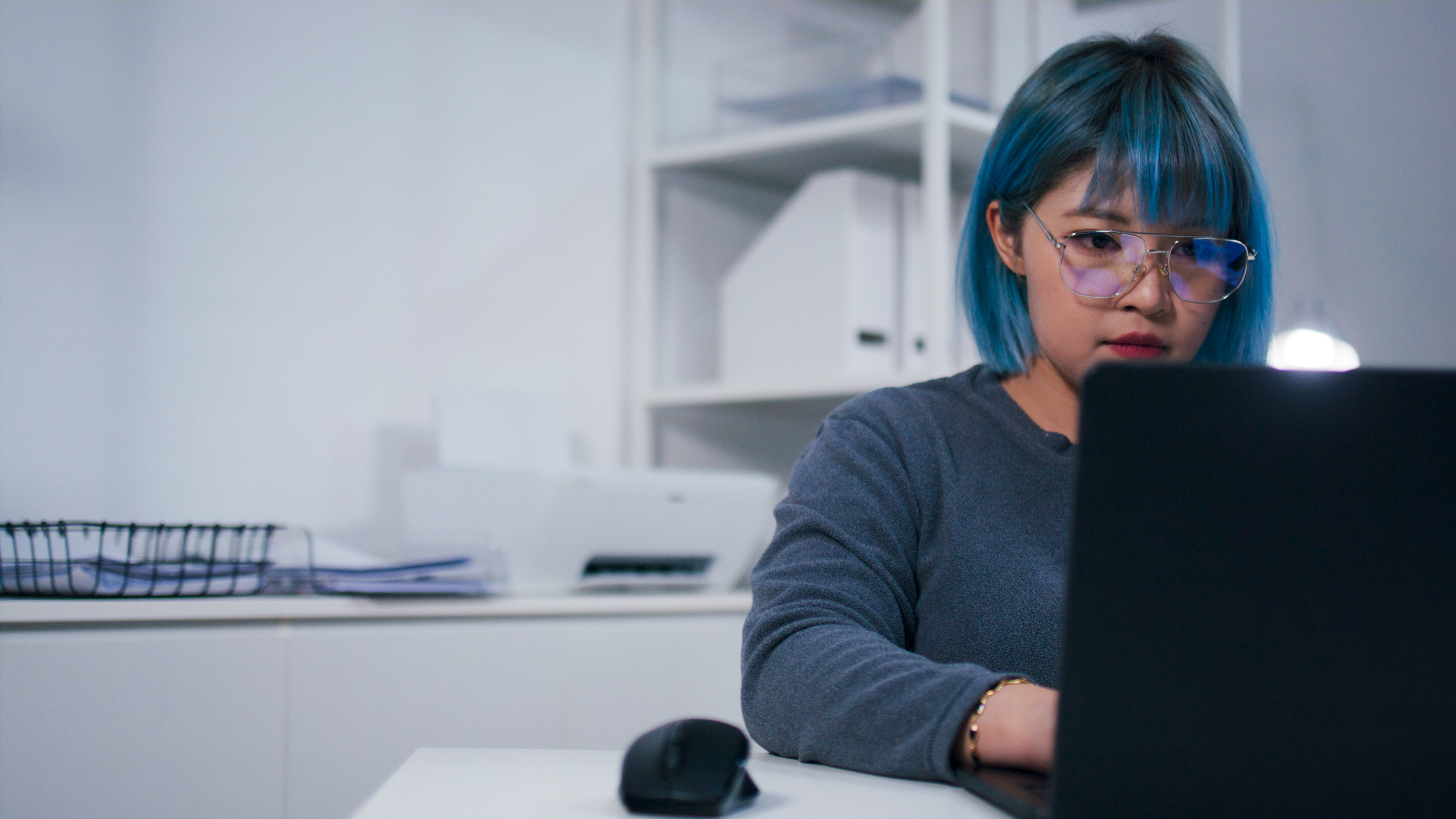 Person with short hair and glasses focuses intently on a laptop, seated at a desk with an organized background