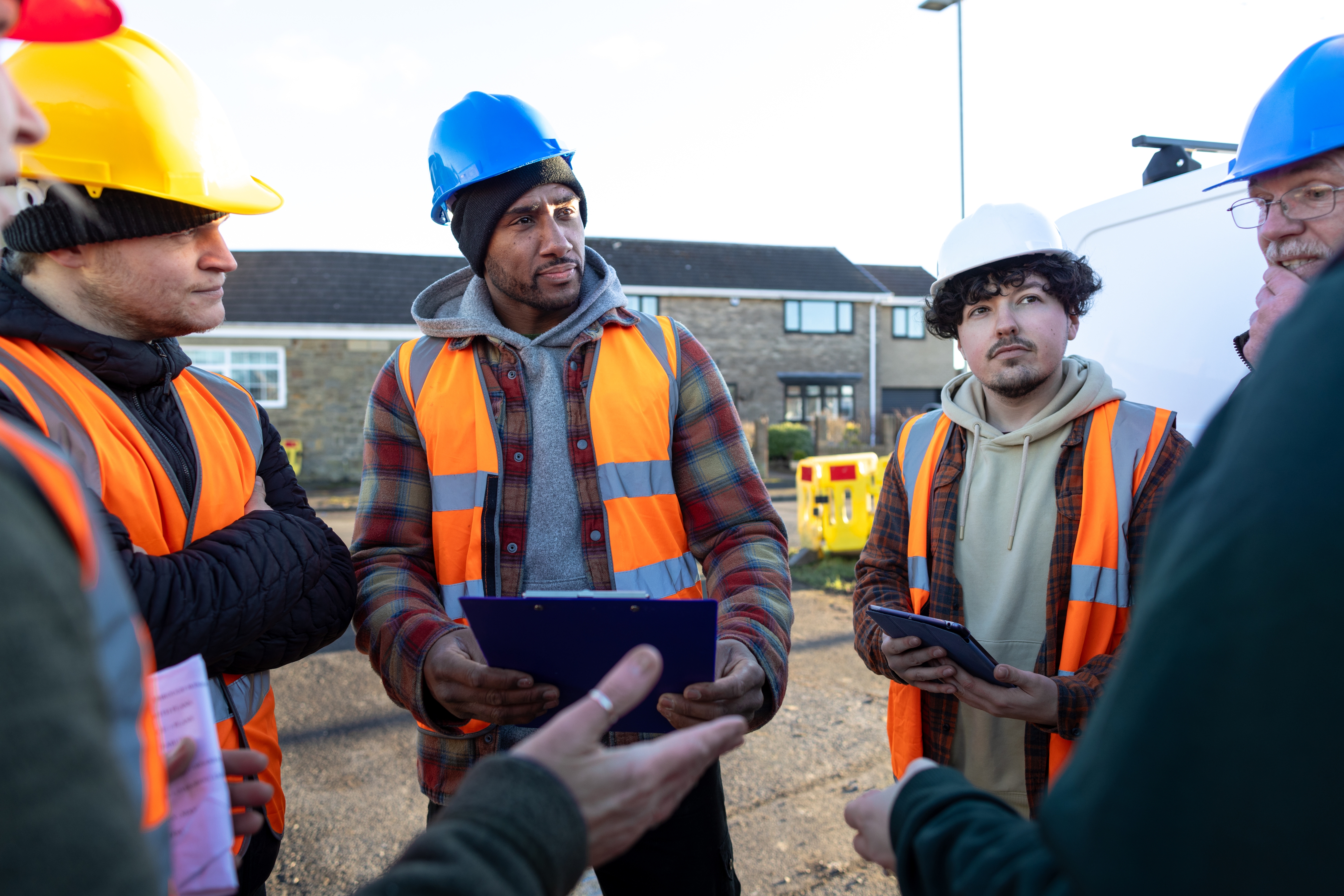 Construction workers wearing safety gear and helmets stand in a group discussing plans outdoors