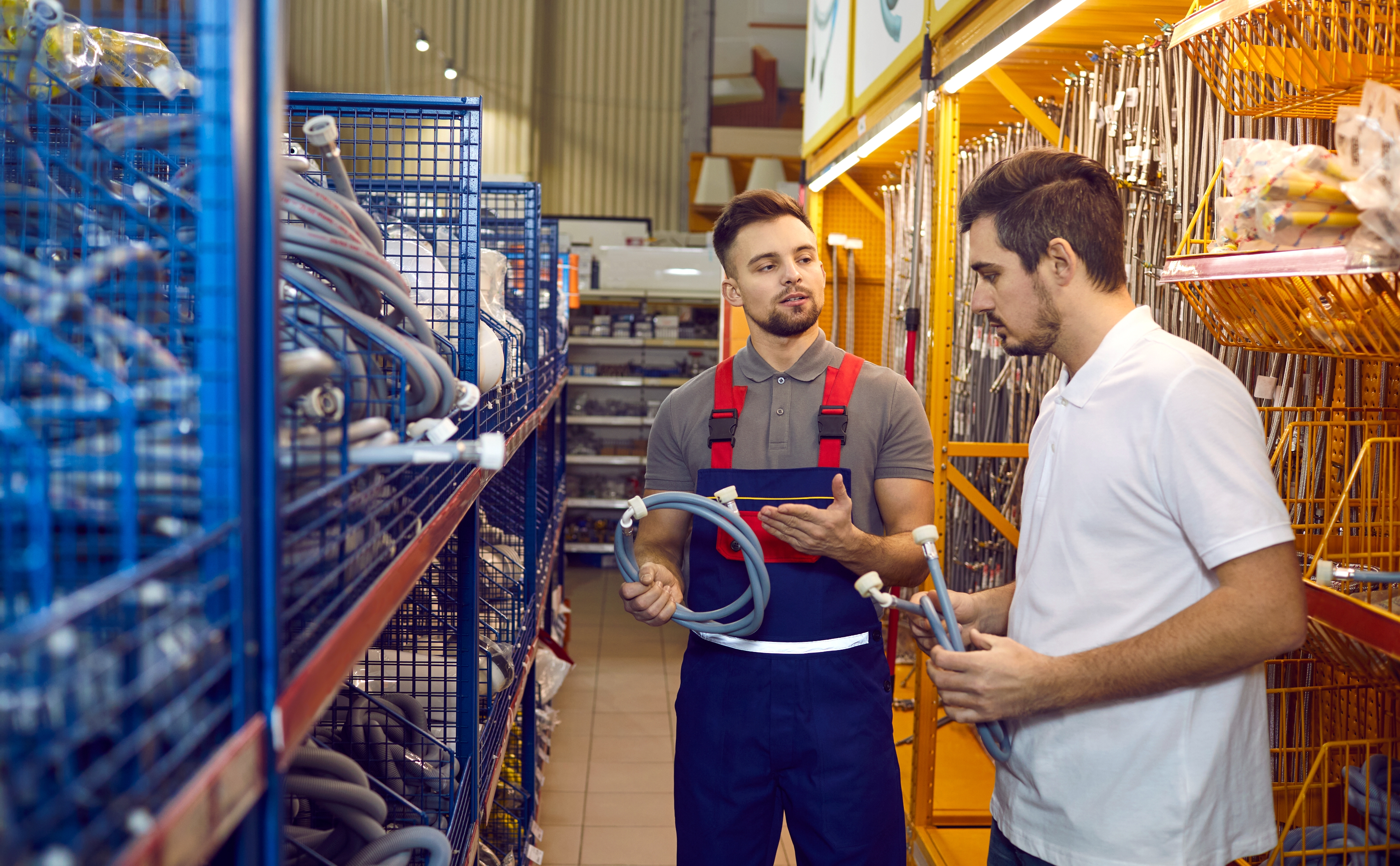 Two men in a store aisle discuss flexible metal tubes. One wears a uniform with red suspenders, indicating his role as an employee or expert