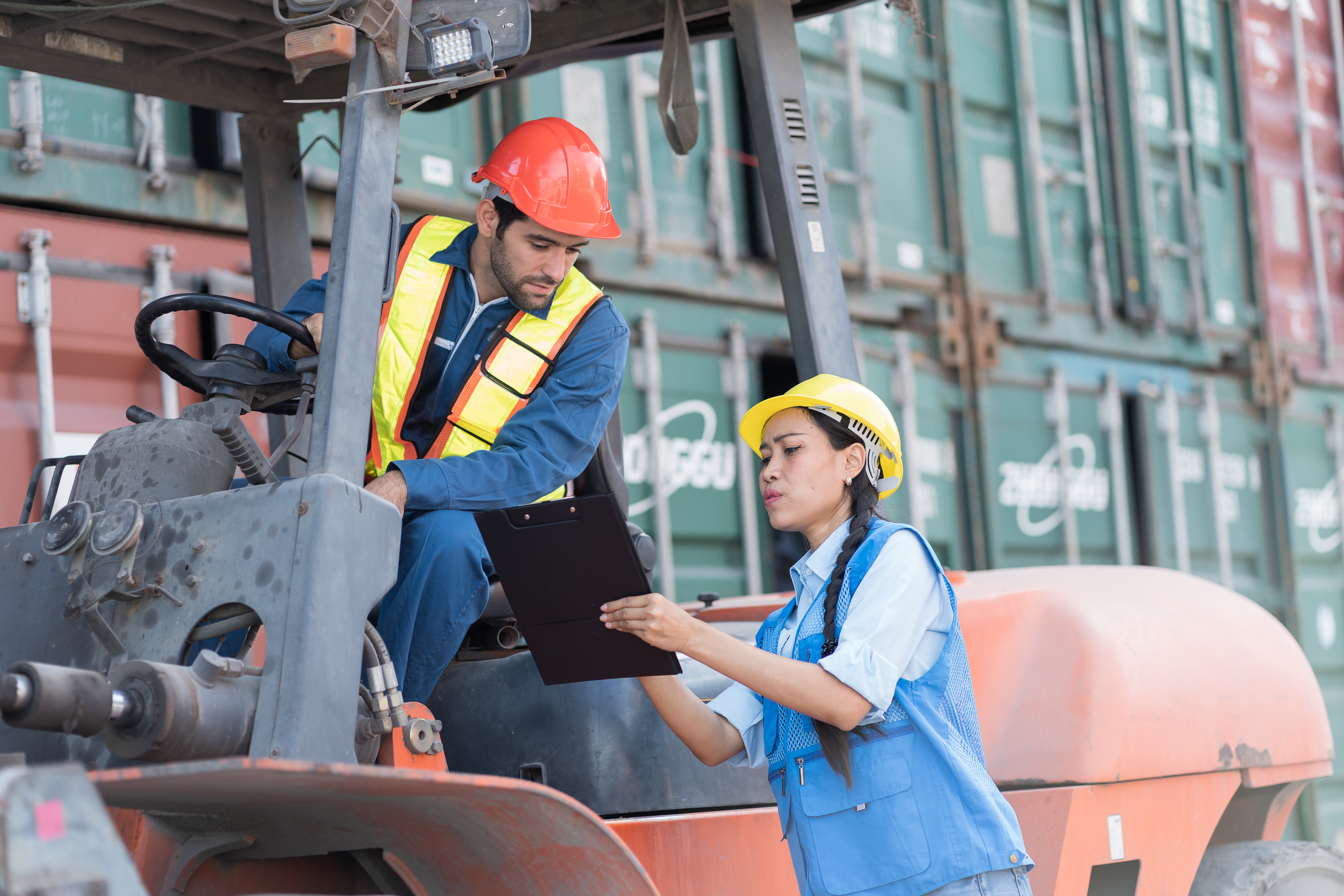 Two industrial workers at a shipping yard; one on a forklift and the other with a clipboard, both wearing safety gear and helmets
