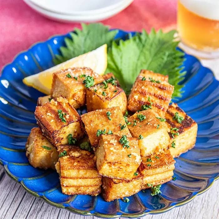 青い皿に盛り付けられたグリルした豆腐の一皿。レモンのくし切りと野菜が添えられている。