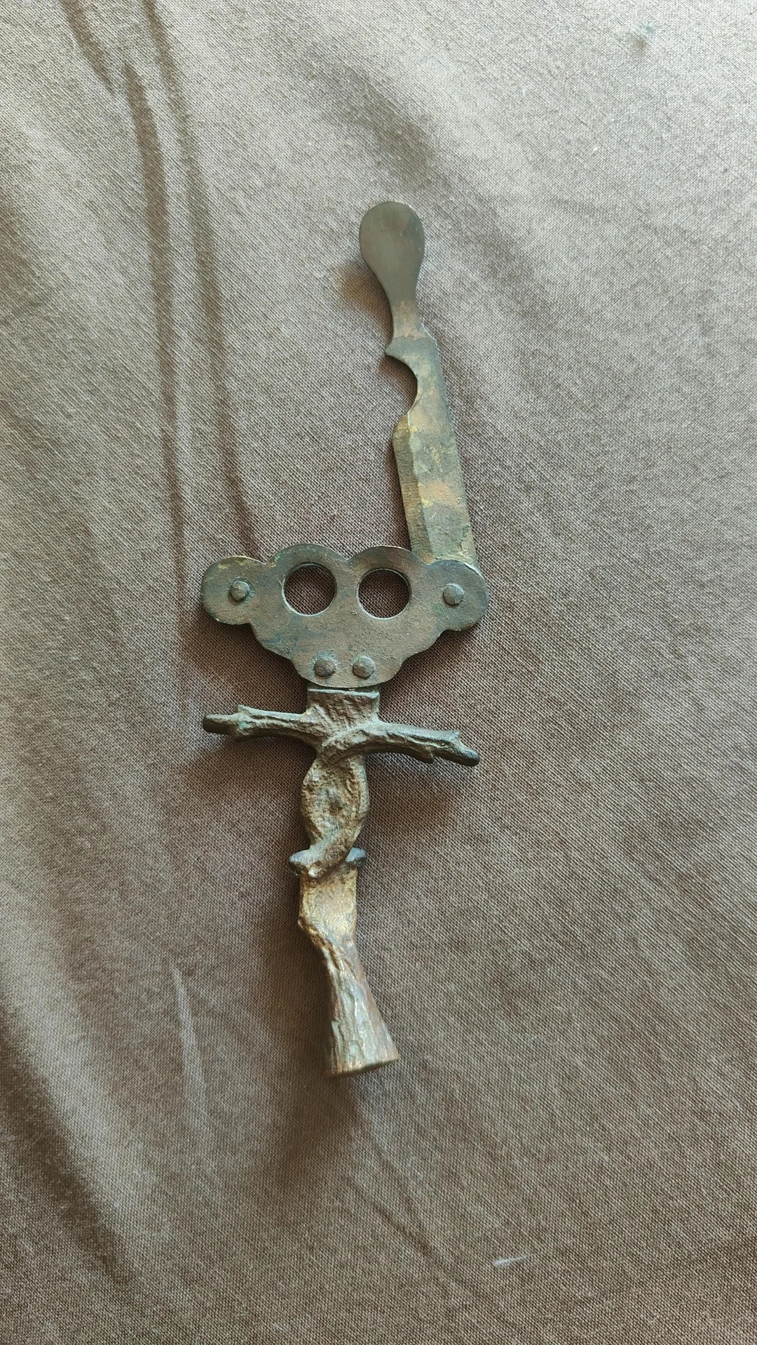 A rustic, handmade metal and wood bottle opener with a unique, twisted handle design, lying on a soft fabric surface