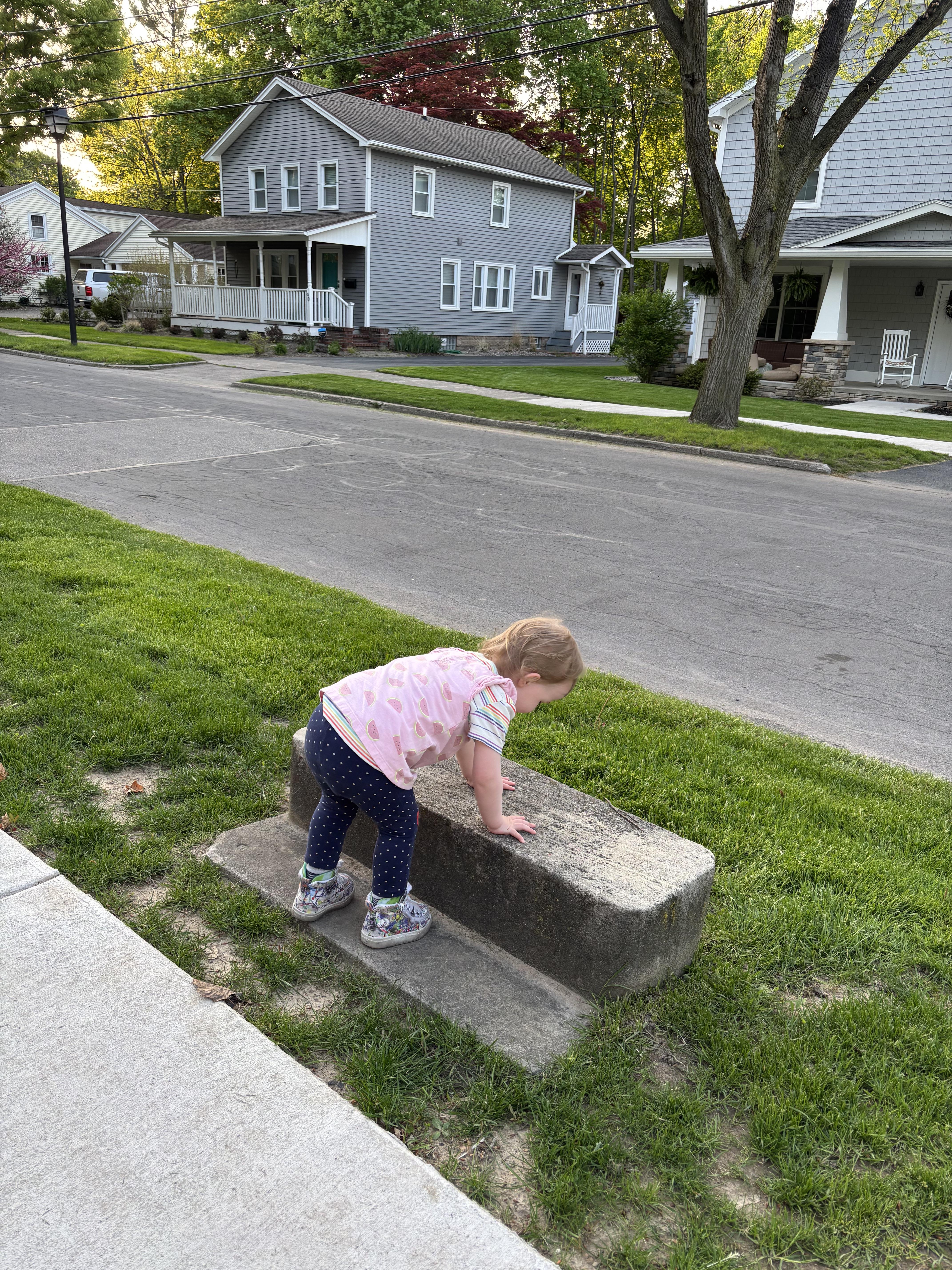 Toddler in casual outfit playing on a concrete block by the sidewalk in a suburban neighborhood