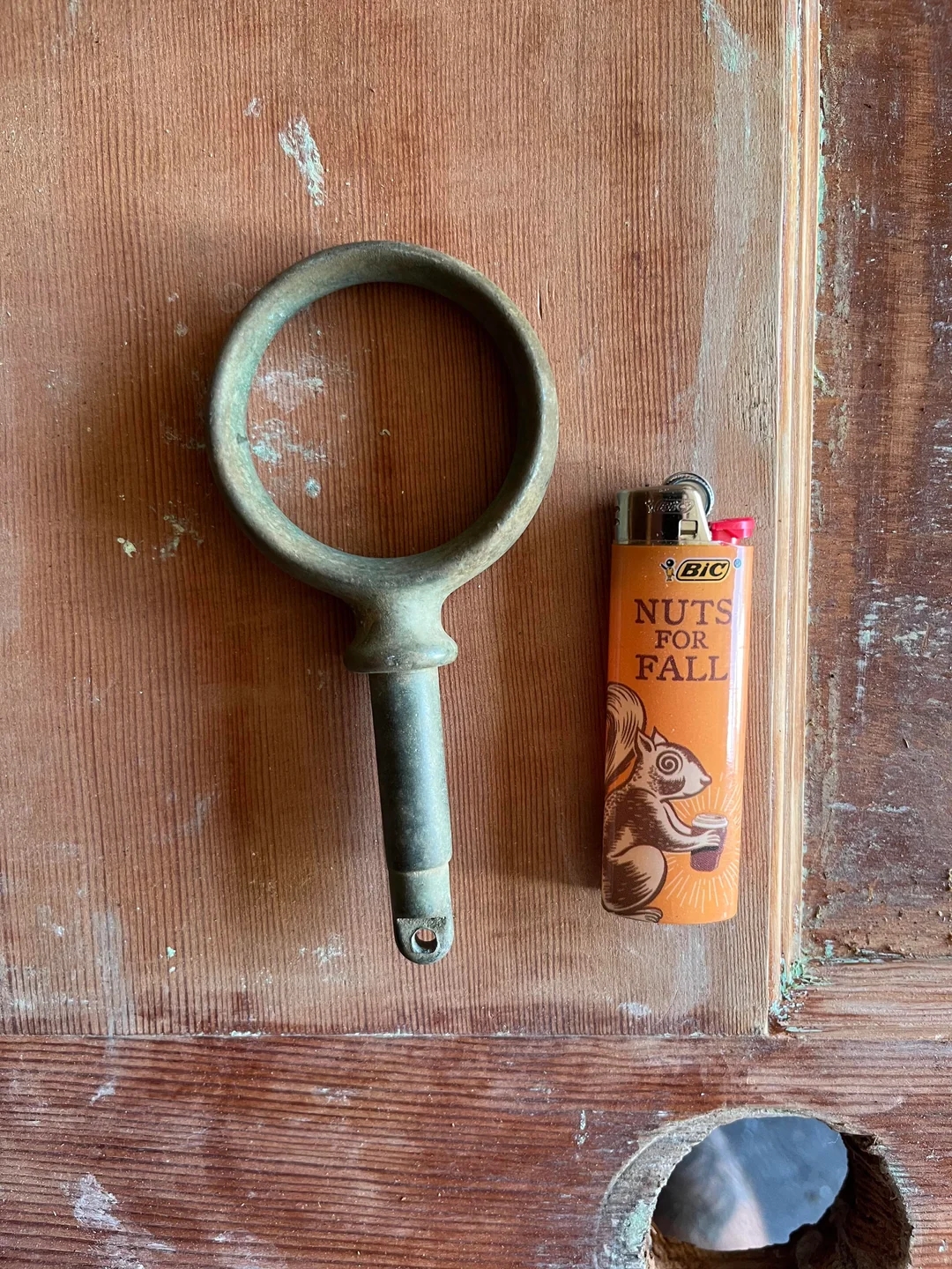 Rusty ring-shaped tool next to an orange lighter with a squirrel image and "Nuts for Fall" text, on a wooden surface