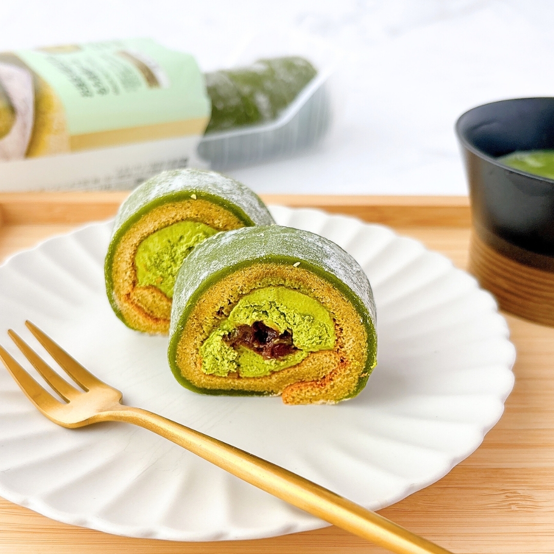 抹茶ロールケーキが白い皿の上に置かれ、金色のフォークが添えられています。隣には飲み物が入った黒いカップがあります。