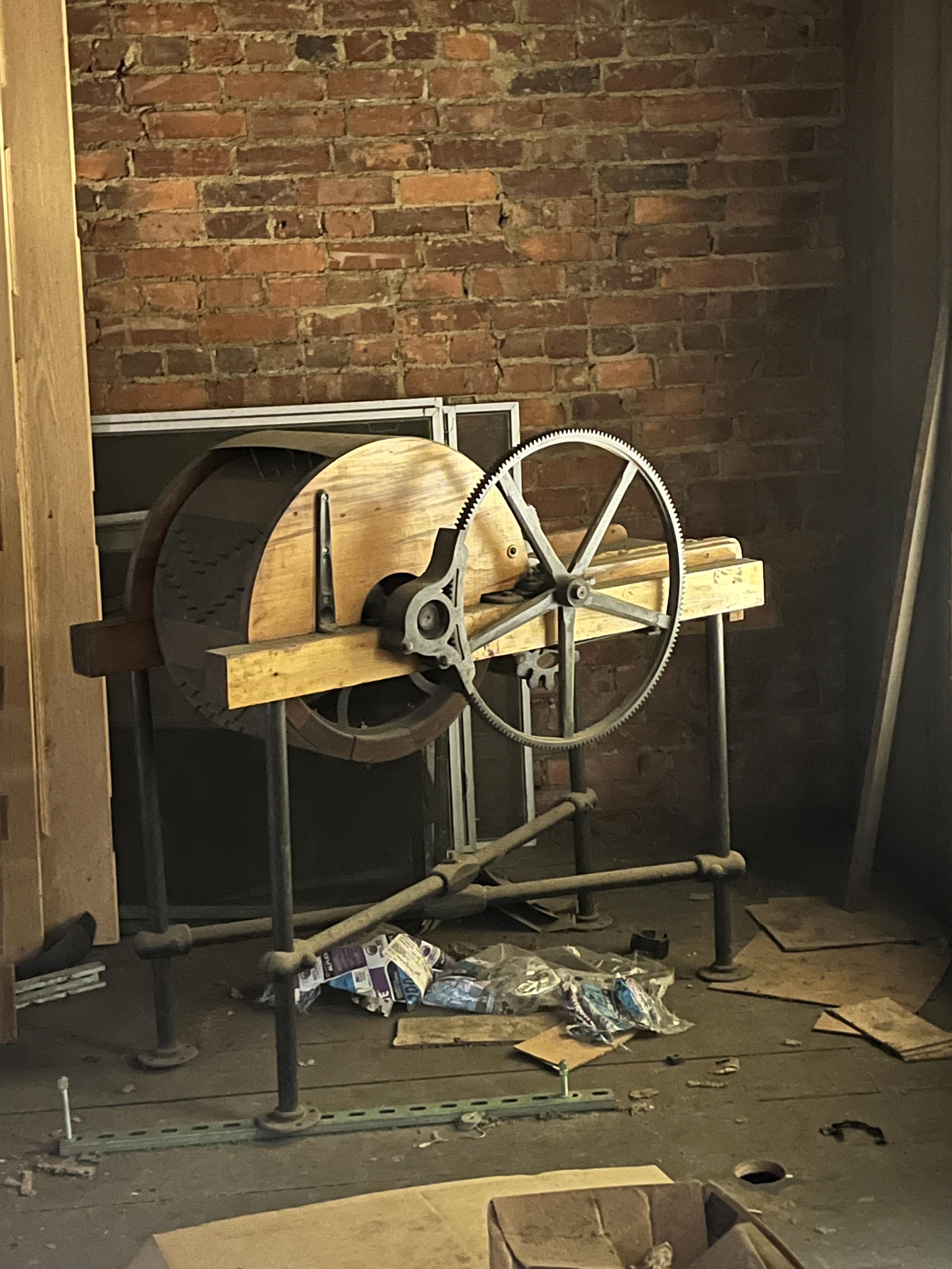 An old wooden and metal machine with gears stands in a rustic room with exposed brick walls and scattered tools