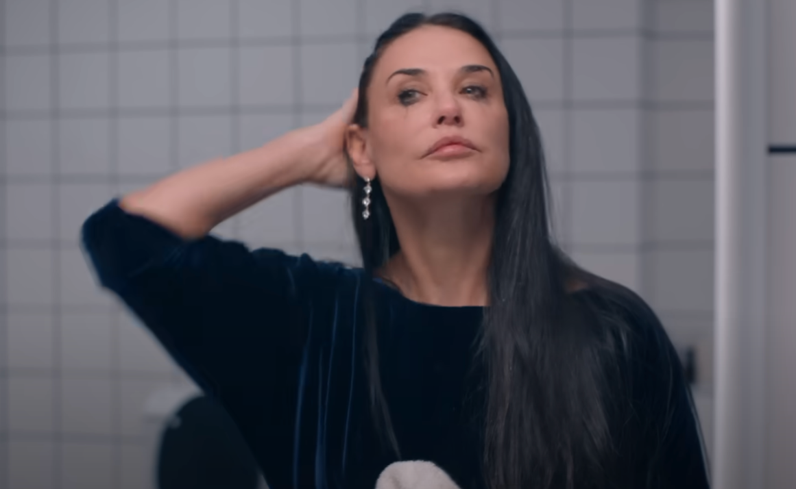 Person with long hair, wearing earrings and a dark top, stands with a hand on their head in a tiled room