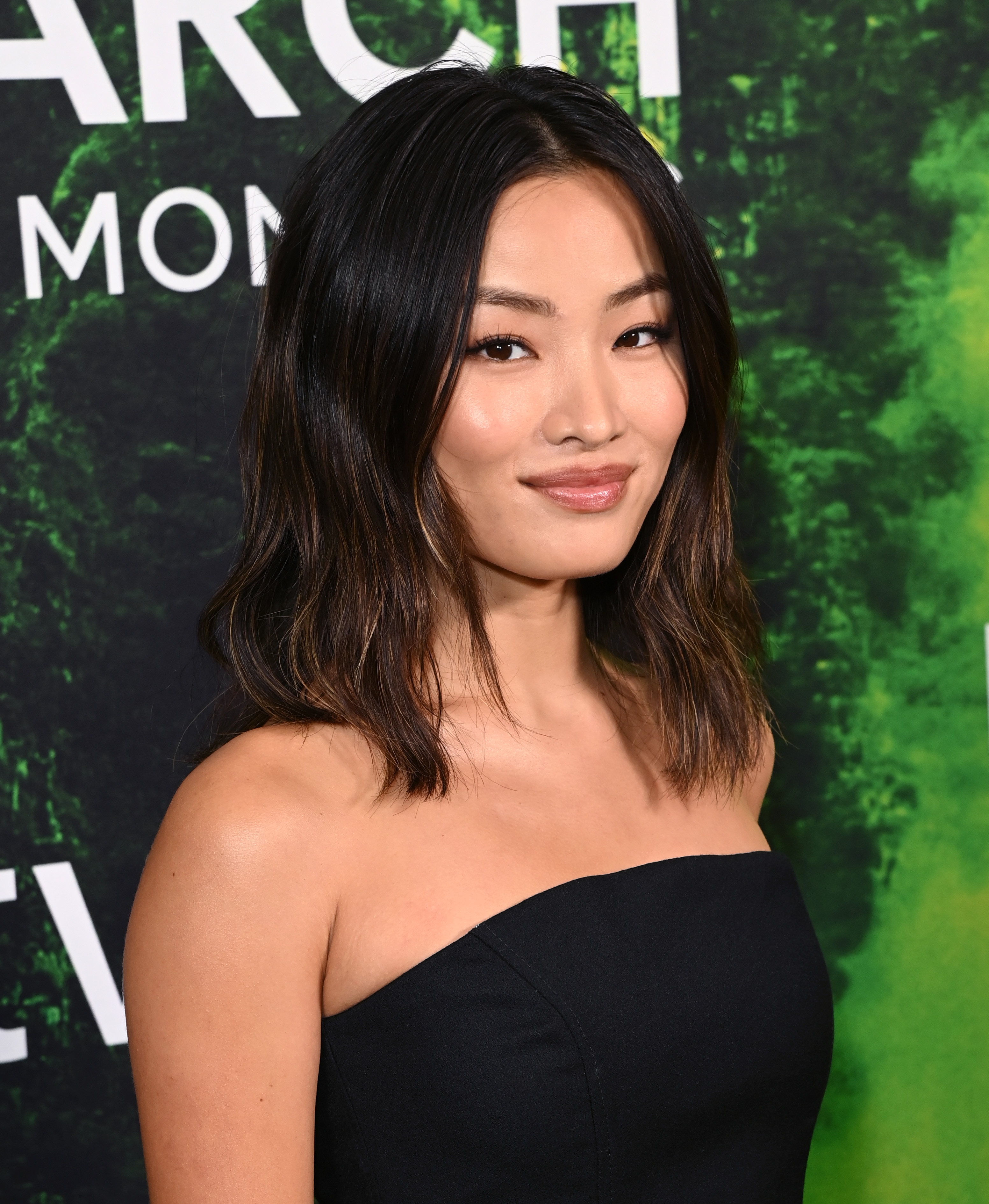 Anna Sawai smiling at a media event, wearing a strapless, elegant dress. Dark, wavy hair frames the face against a green-hued background