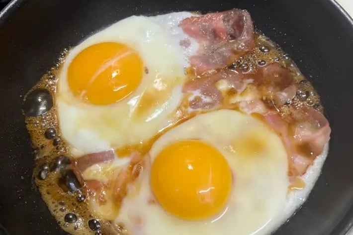 フライパンでベーコンと目玉焼き2つが焼かれている様子。