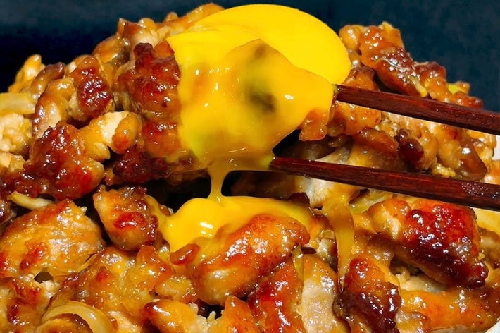焼き肉丼の上に半熟卵がのせられ、割られた黄身が流れ出している様子。美味しそうな料理が強調されている。