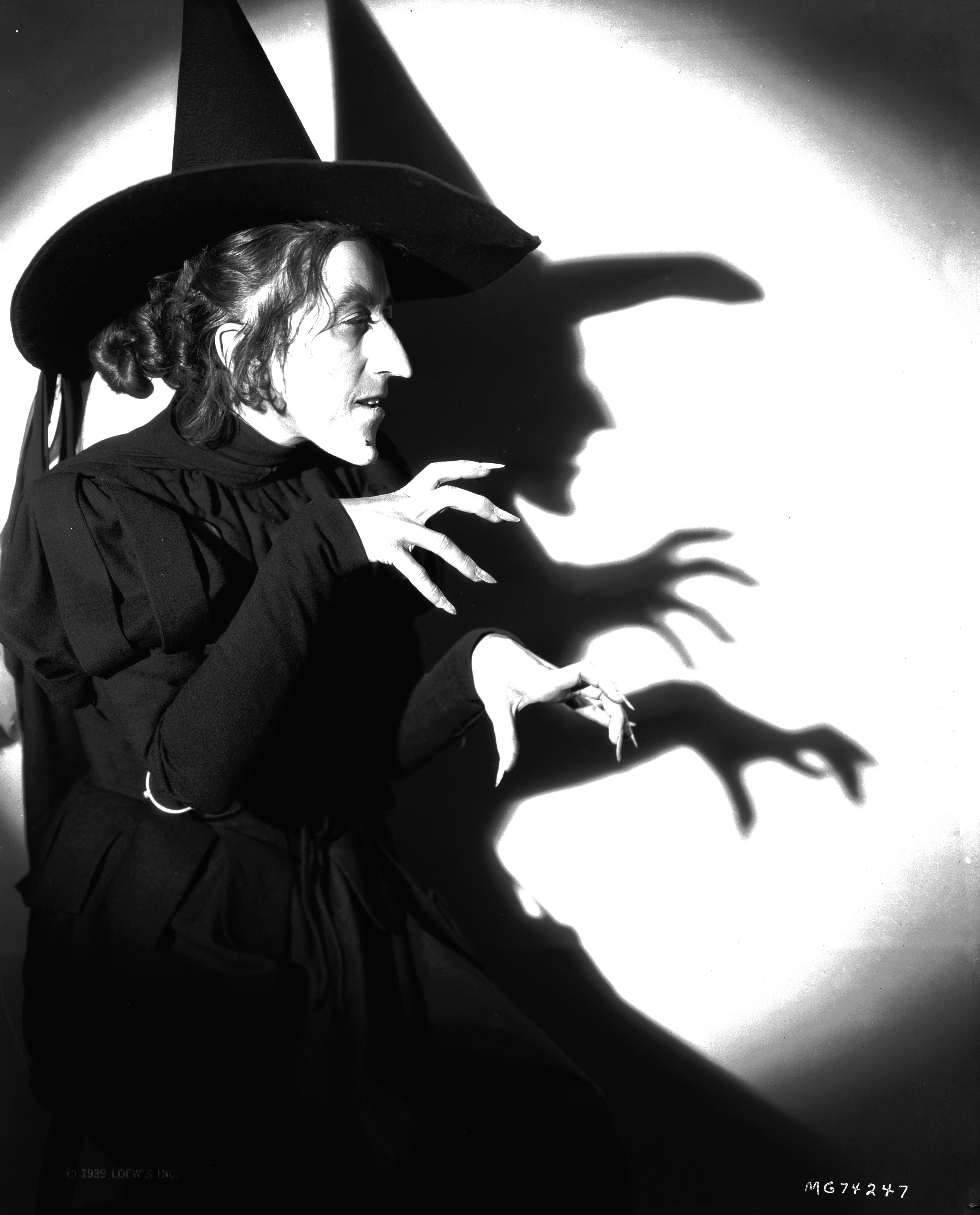 Person in a witch costume with a pointed hat and long cloak poses dramatically, casting a large shadow on the wall