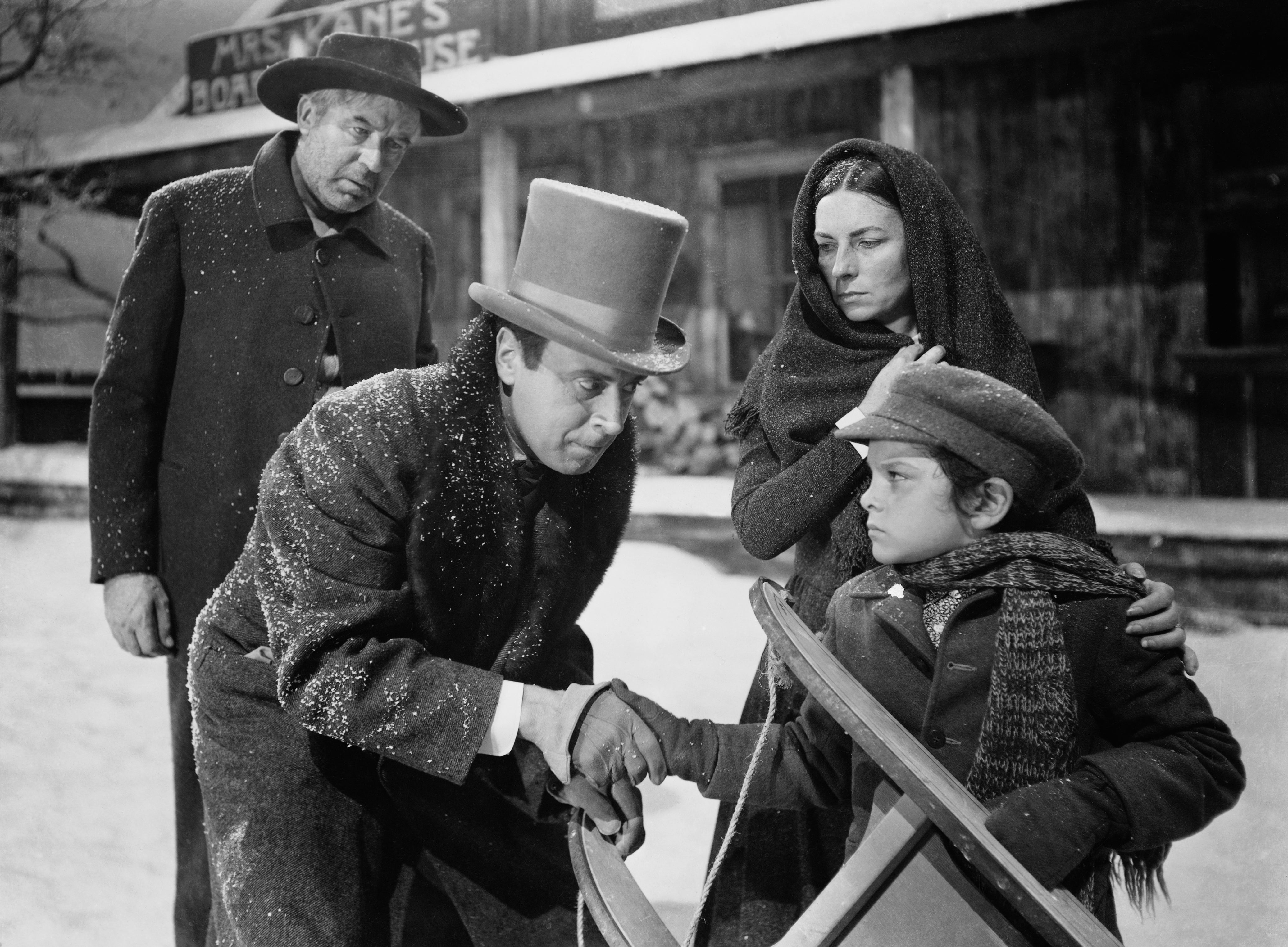Period film scene with four individuals in winter attire. A man in a top hat shakes hands with a boy, while two others watch in a snowy setting