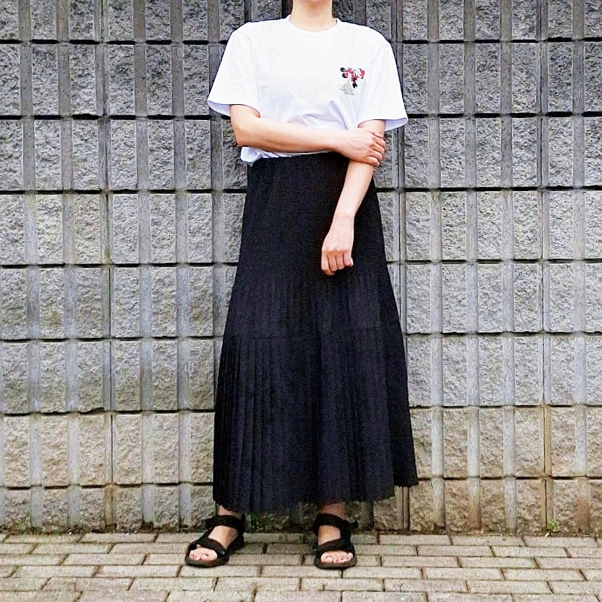 女性が壁の前で白いTシャツと黒いプリーツスカートを着て立っている。