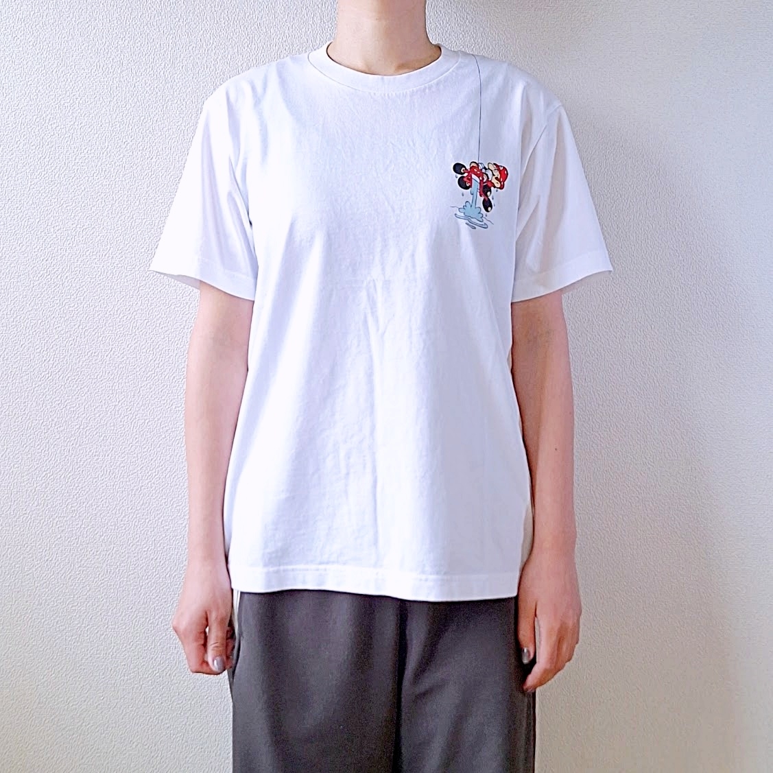 人物が白いTシャツとシンプルなパンツを着て立っている。Tシャツには小さなデザインがある。