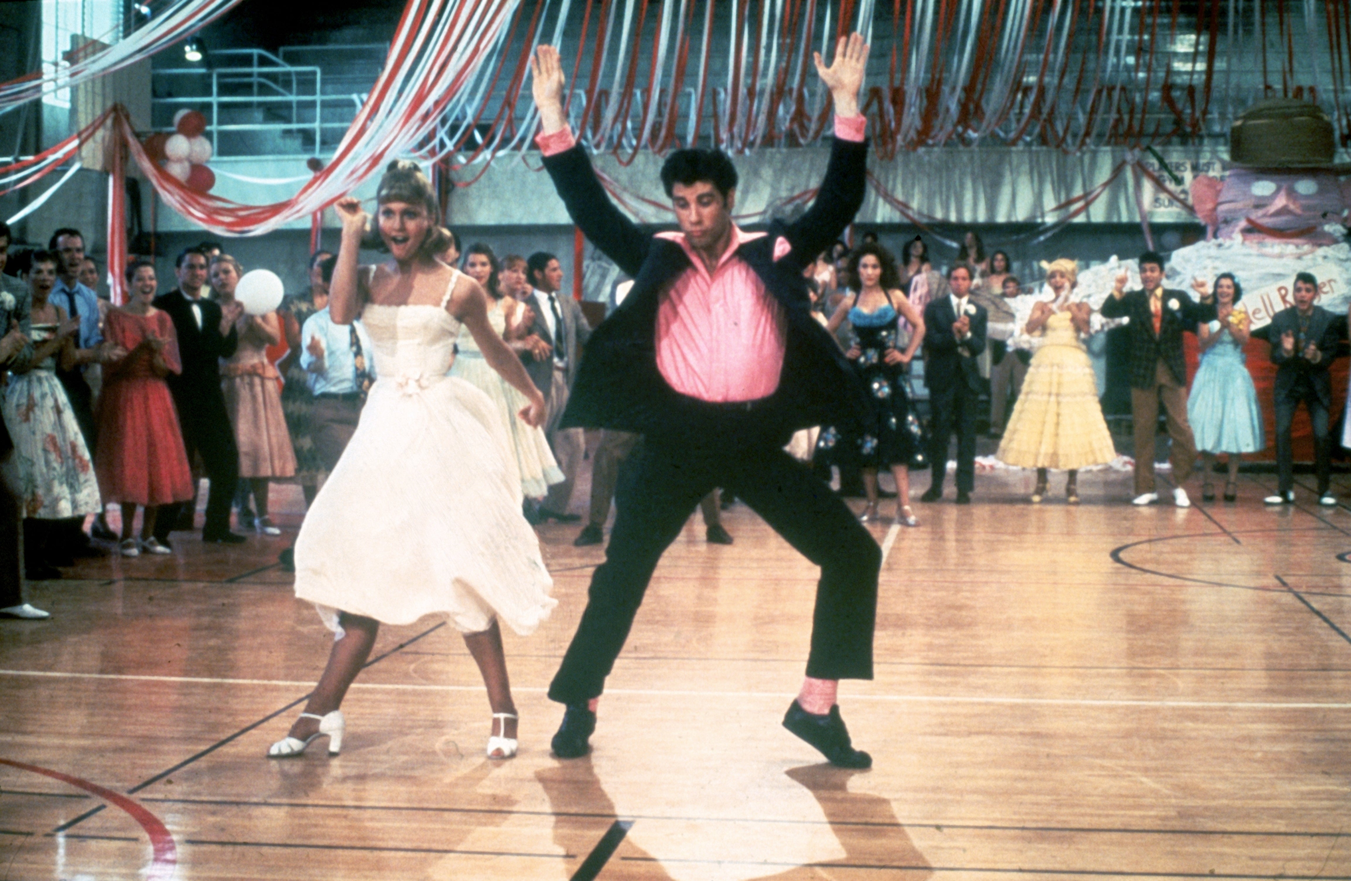 A lively dance scene with two performers dressed in 1950s-style outfits, dancing energetically in a gymnasium with a cheering crowd