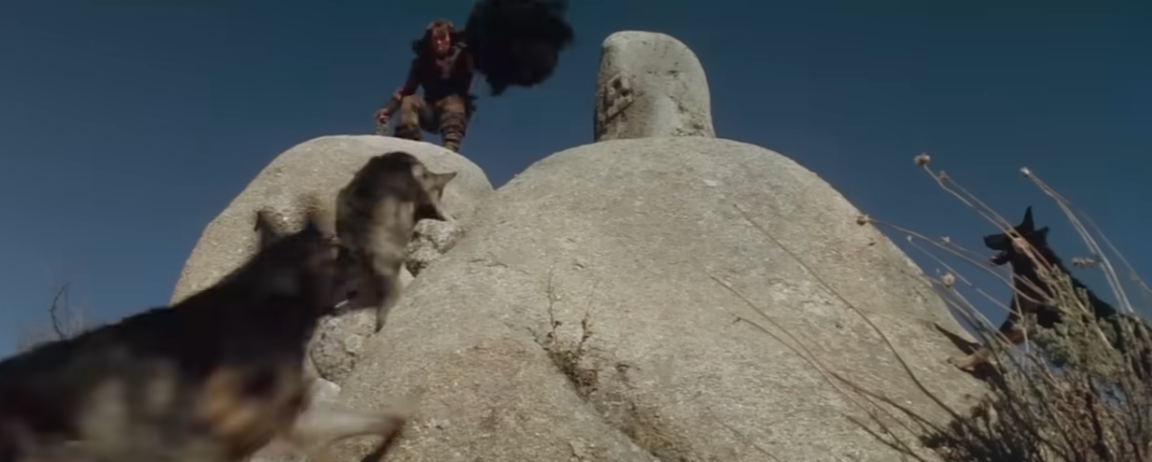 Person standing on rocks with two wolves approaching below, in a dramatic outdoor scene