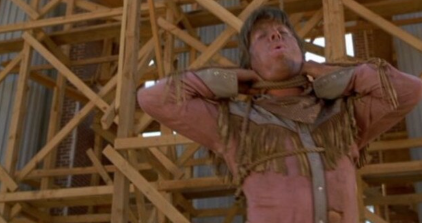 Person in dusty, fringed Western-style outfit dramatically poses in front of wooden scaffolding