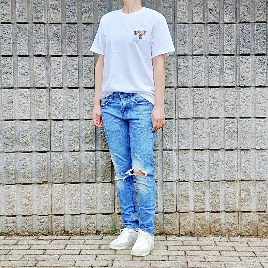 白いTシャツにダメージジーンズ、スニーカーを履いた立っている人物。背景は石の壁。