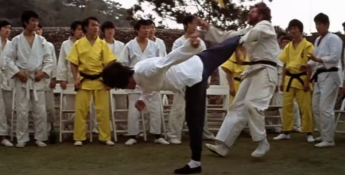 Martial artists in white and yellow uniforms watch a high kick demonstration in an outdoor setting