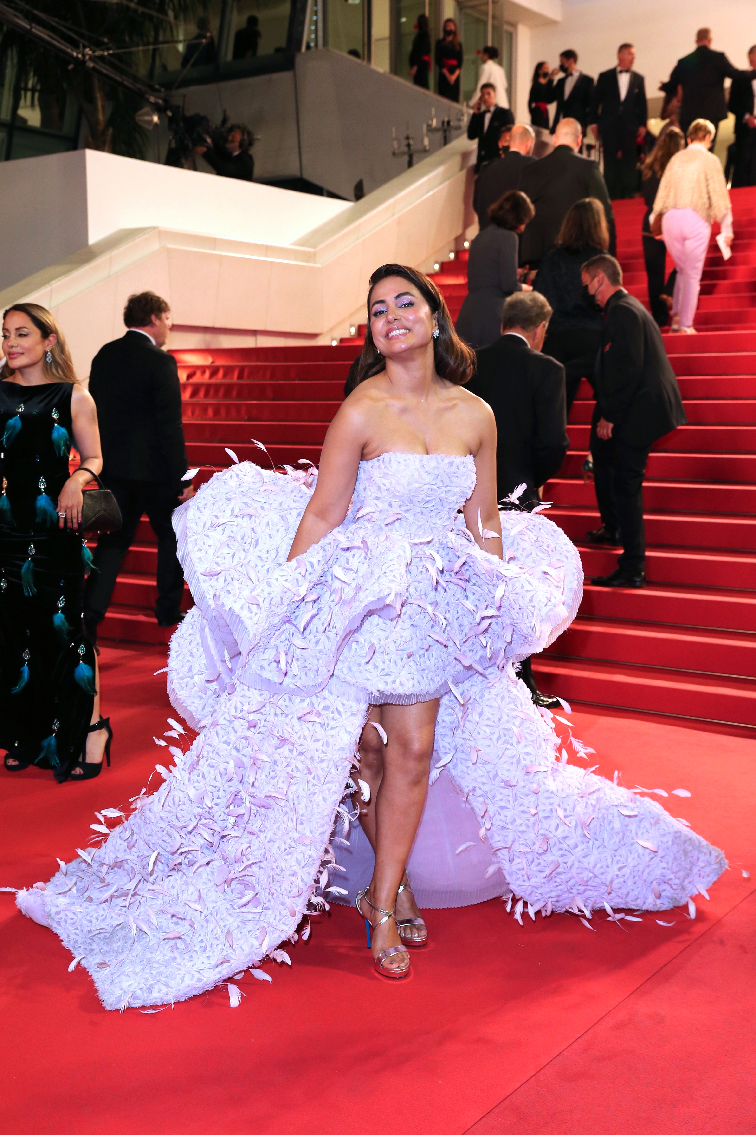 Celebrity in an elegant, voluminous gown with ruffled details poses on a red carpet with people and photographers in the background