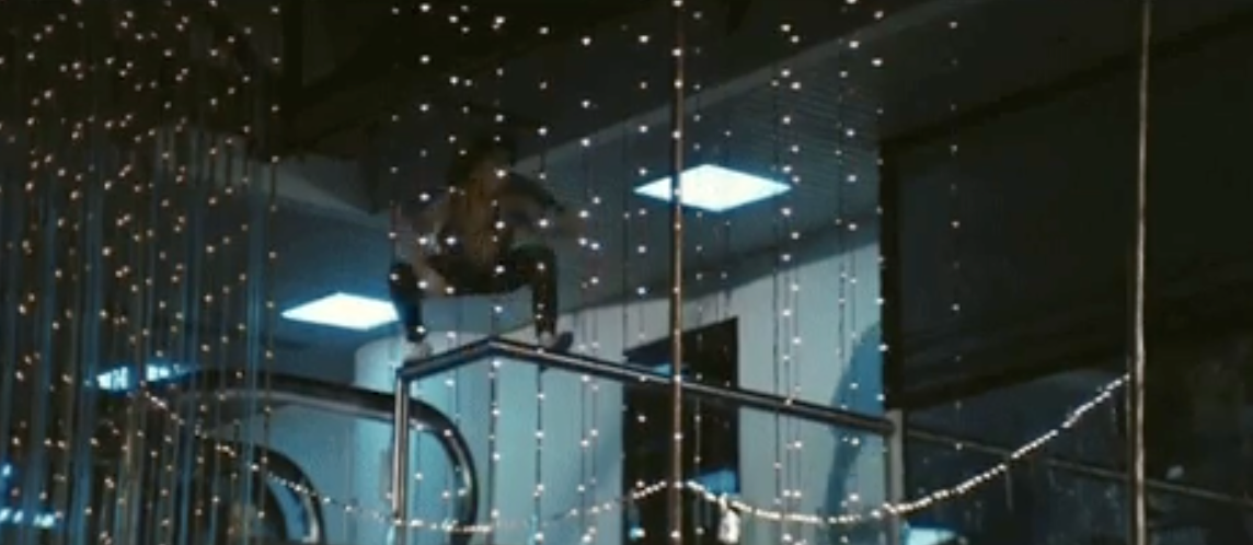 A person climbs a railing in a dimly lit area decorated with hanging lights, resembling a festive or holiday setting