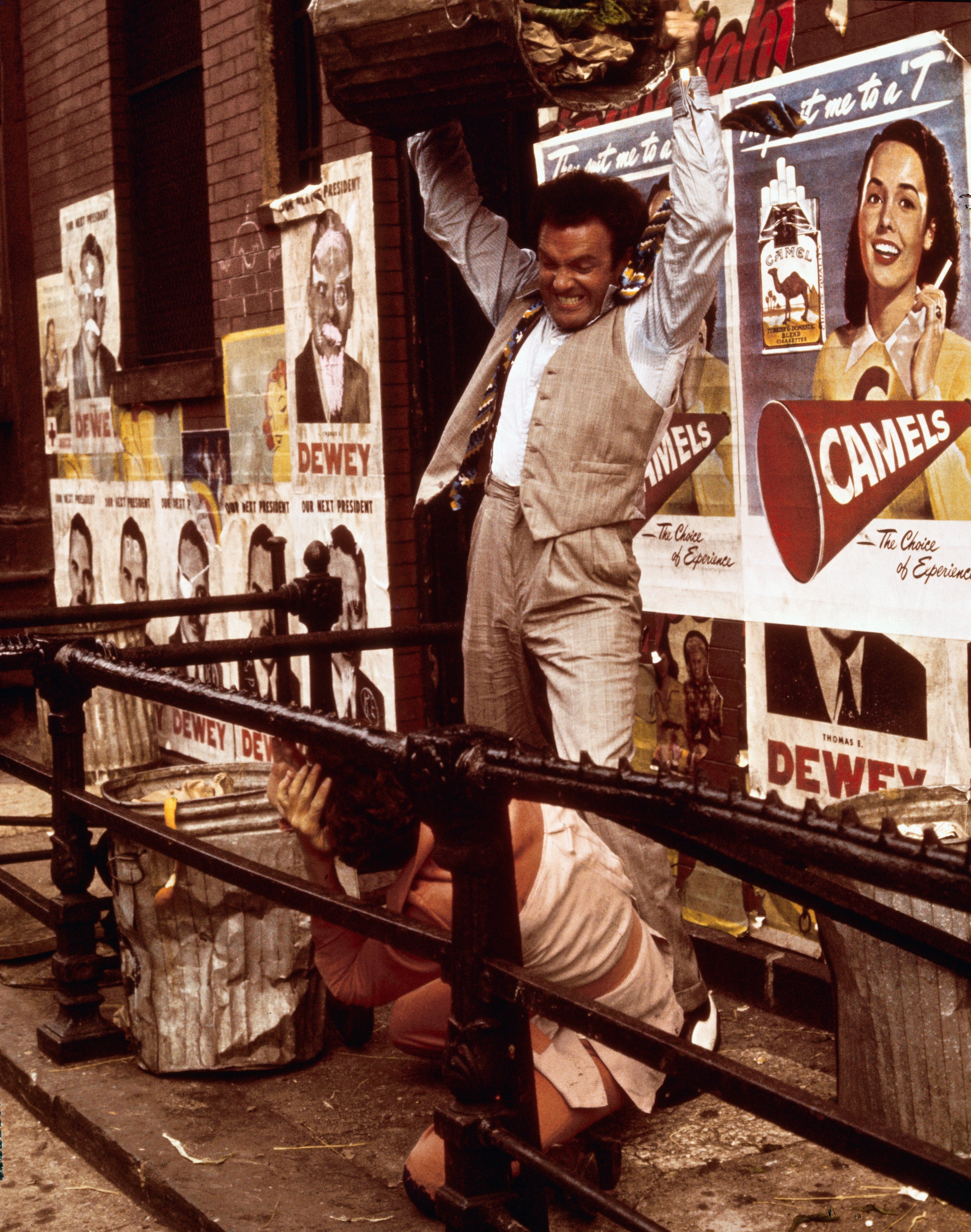 A person in a vintage suit holds a crate above someone crouched behind a railing, against a wall with retro advertisement posters