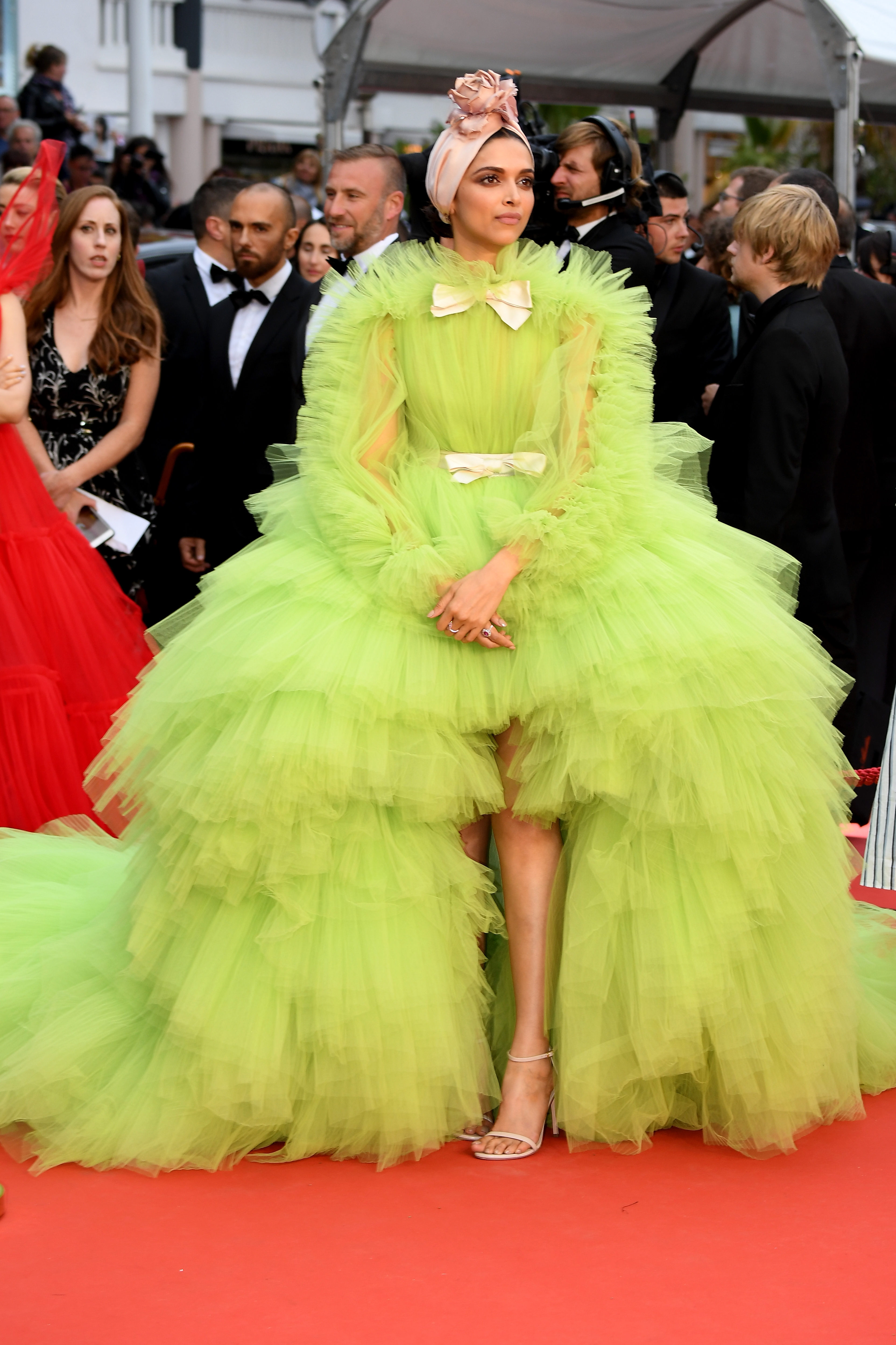 Person on red carpet in voluminous, tiered gown with ruffled layers, styled with a headpiece and strappy heels. Other attendees visible in background
