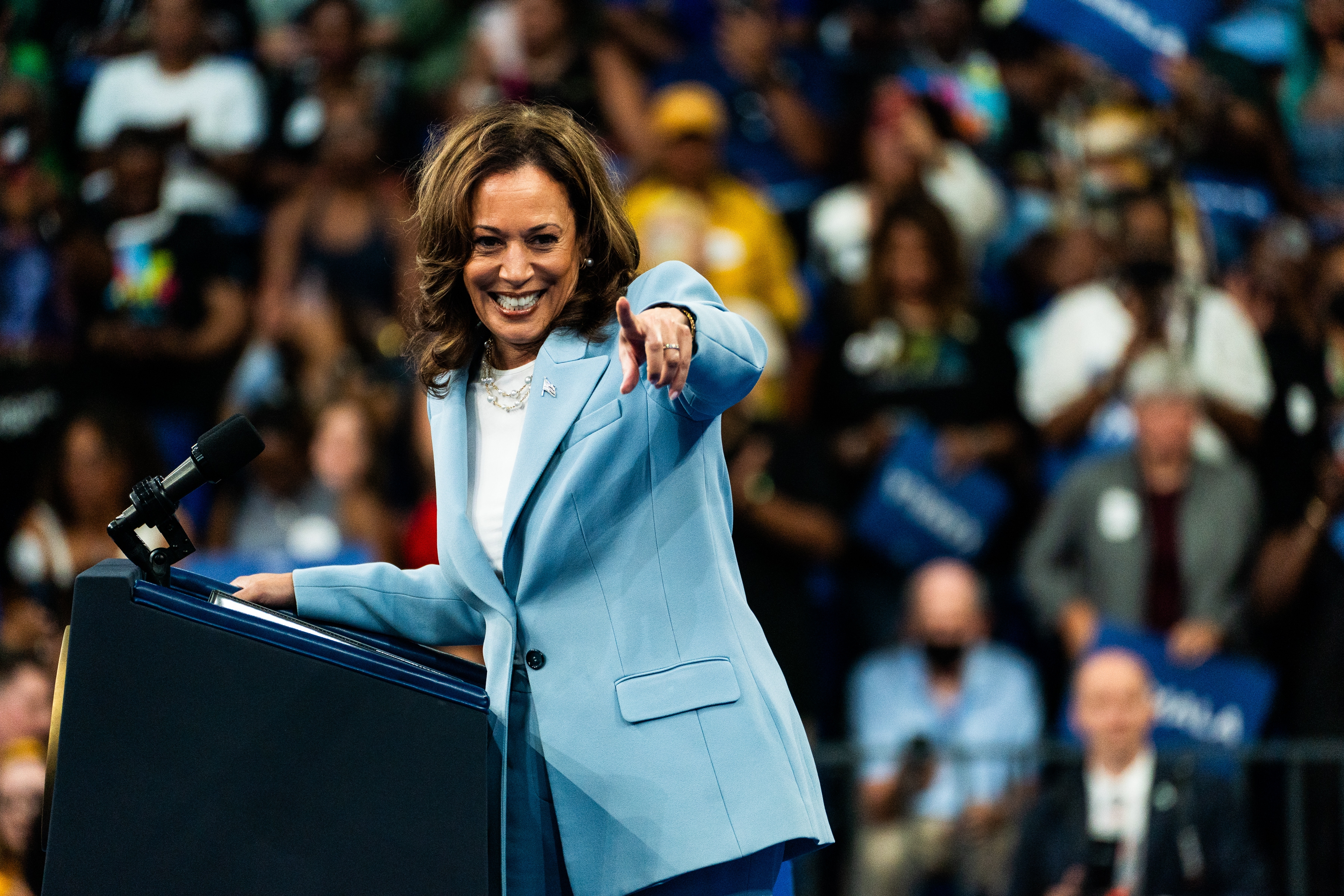 Kamala Harris smiling and pointing while speaking at a podium surrounded by a cheering crowd