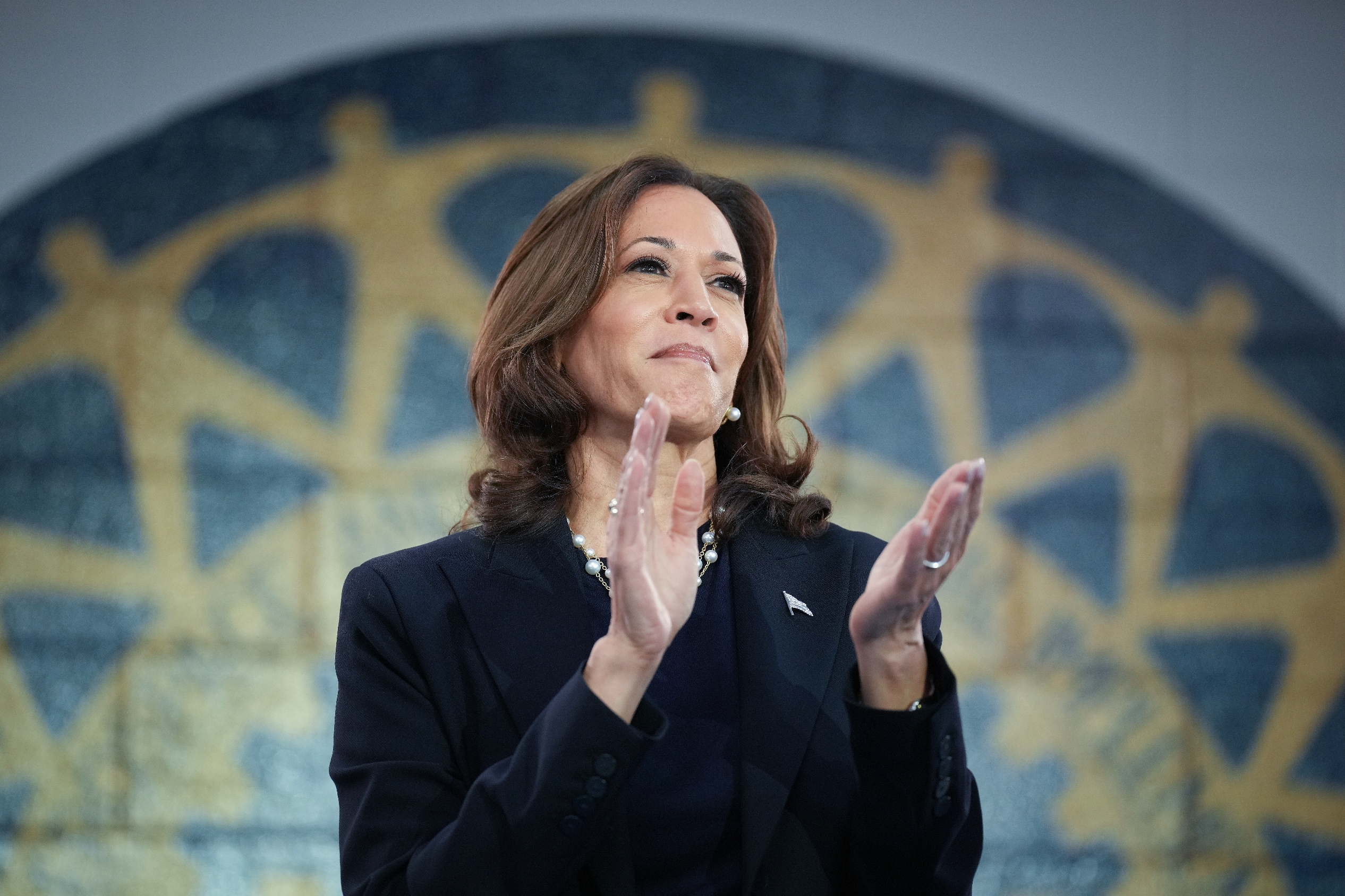 Kamala Harris in a dark suit claps at an event, standing before a large artistic backdrop