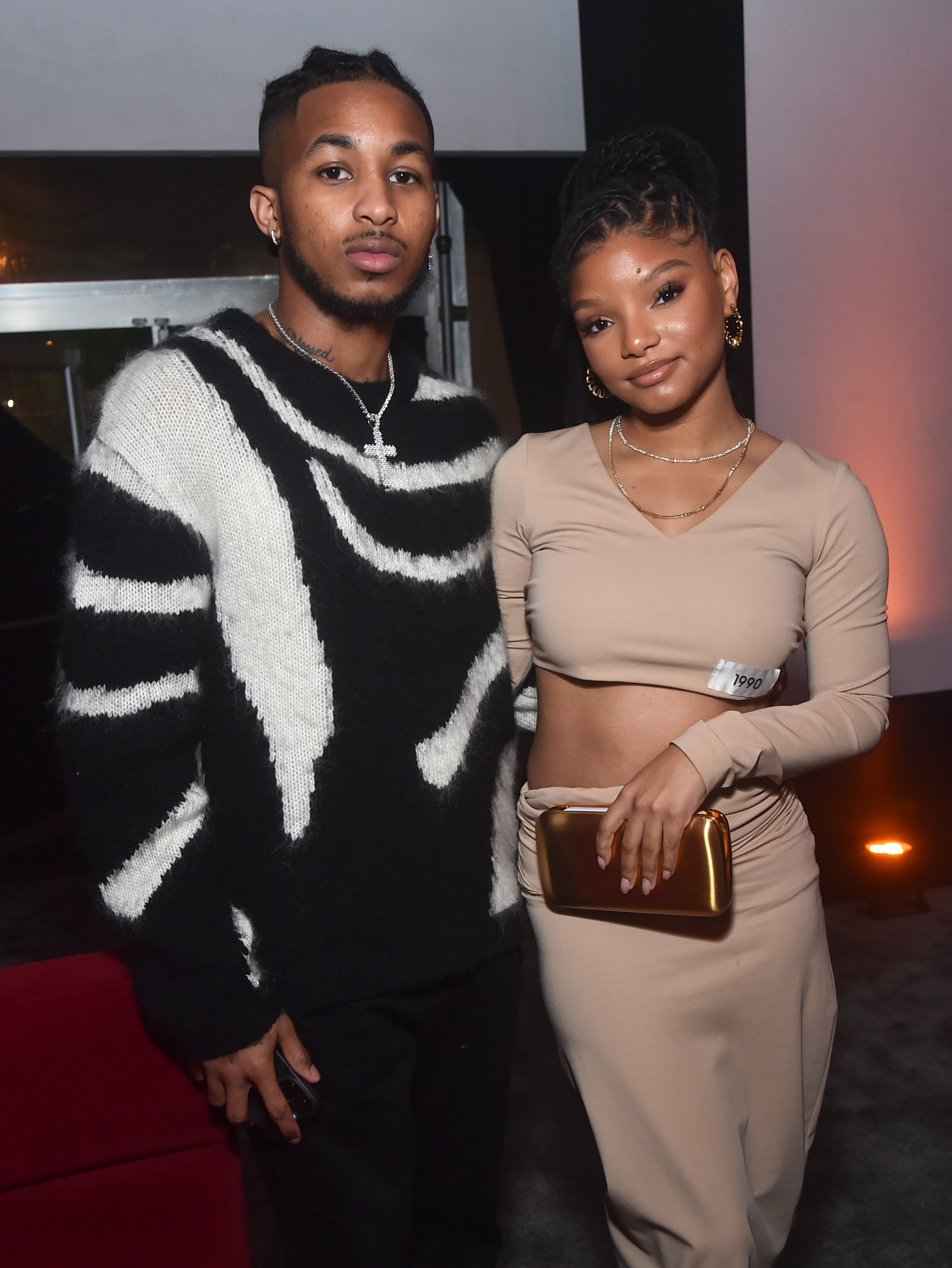 Two celebrities pose together. The woman wears a stylish crop top and skirt, holding a clutch. The man wears a bold patterned sweater
