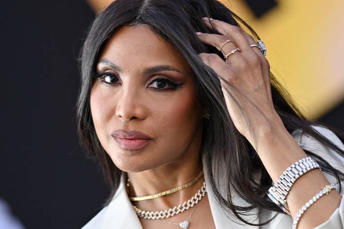 Toni Braxton wearing a blazer and layered necklaces, gently touching her hair, at a celebrity event