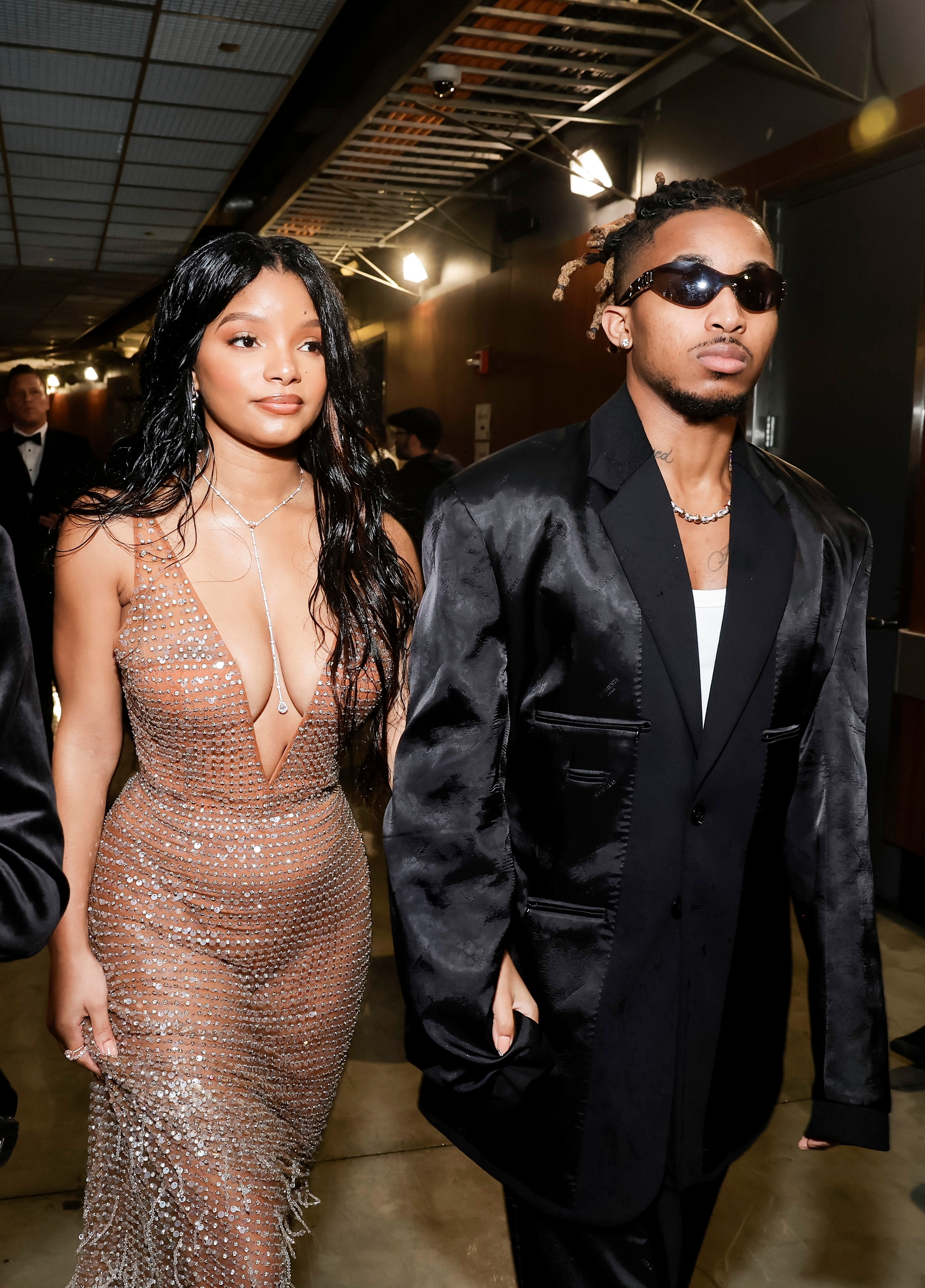 Two public figures are at a formal event. One wears a glittering, form-fitting gown; the other wears a sleek suit with sunglasses