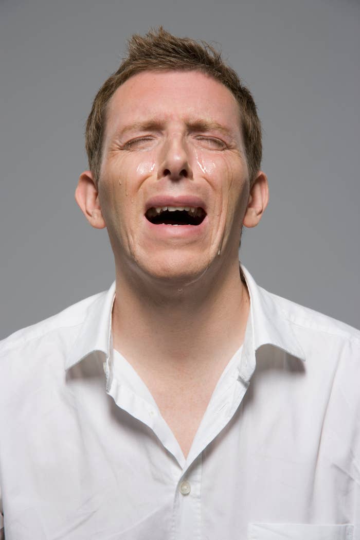 A person is seen crying with eyes closed, tears on their cheeks, wearing a button-up shirt. Their expression conveys sadness or distress