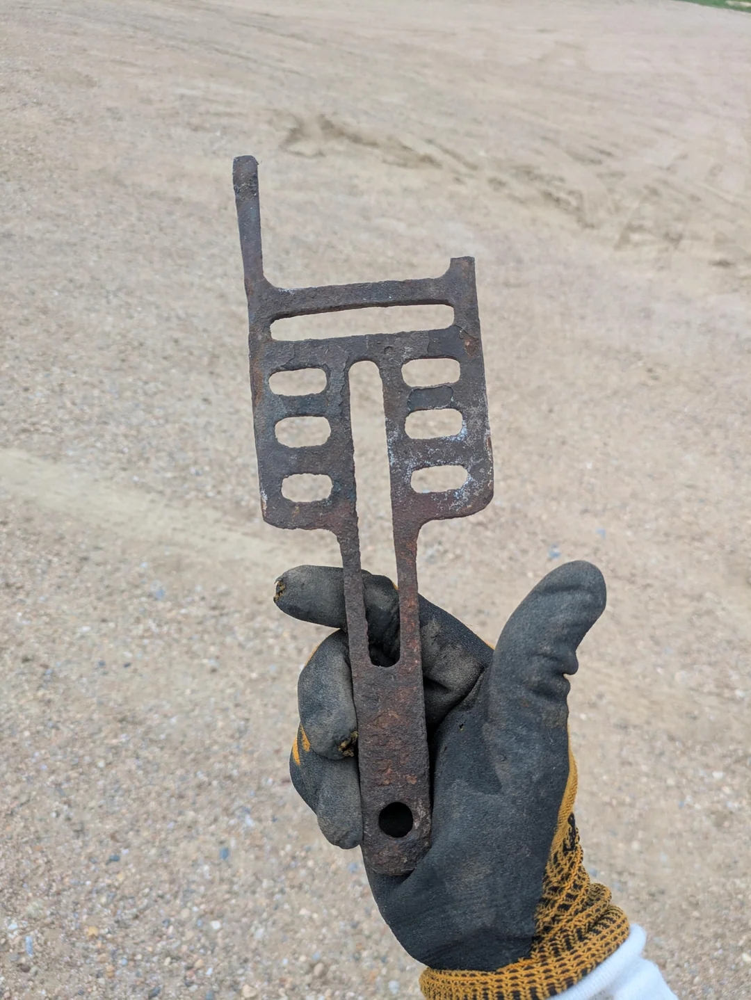 A gloved hand holds a rusty, perforated metal object with a flat handle, against a gravel or sandy background