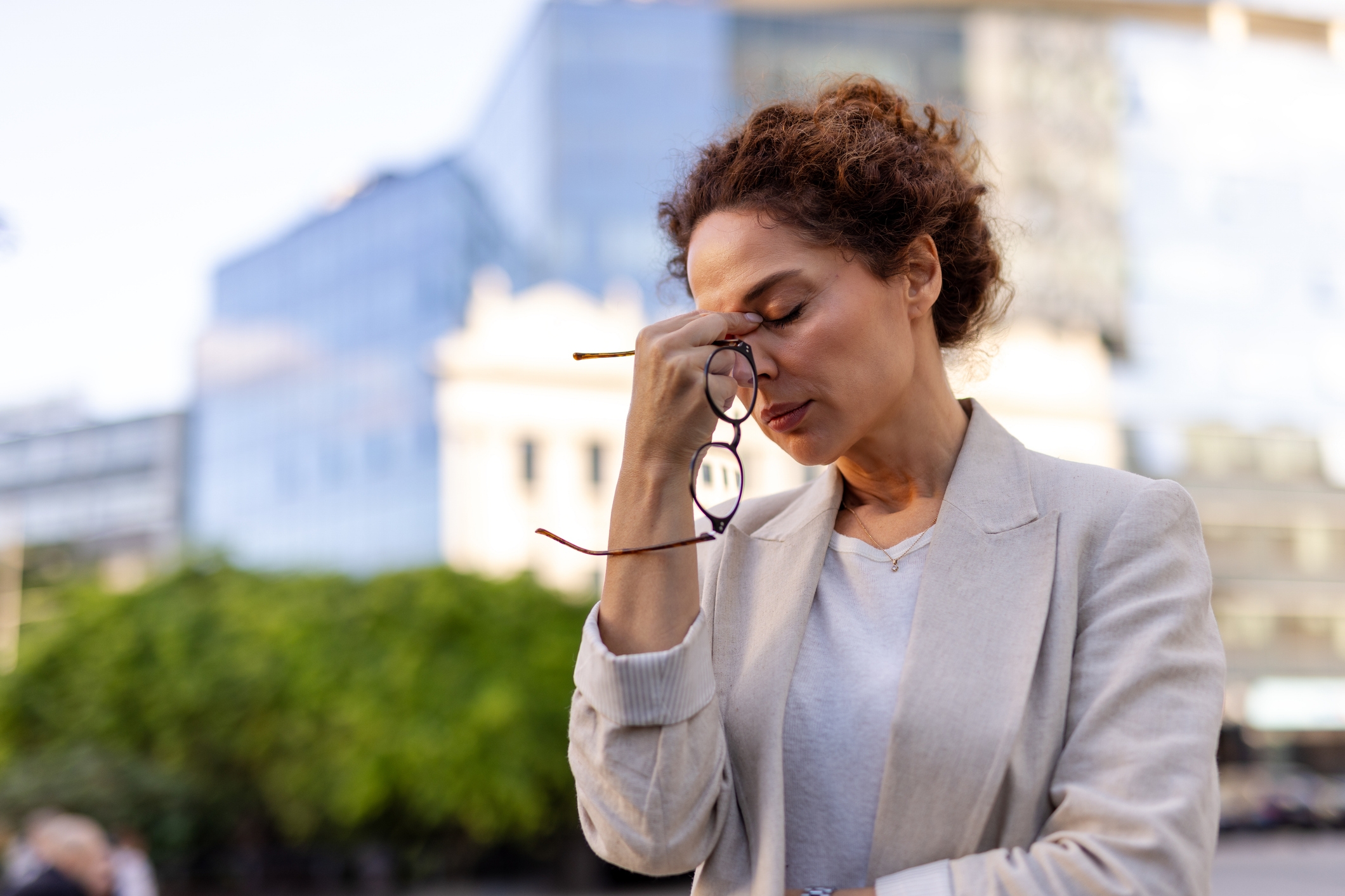 A person stands outdoors, holding glasses, appearing thoughtful or stressed, with an urban background