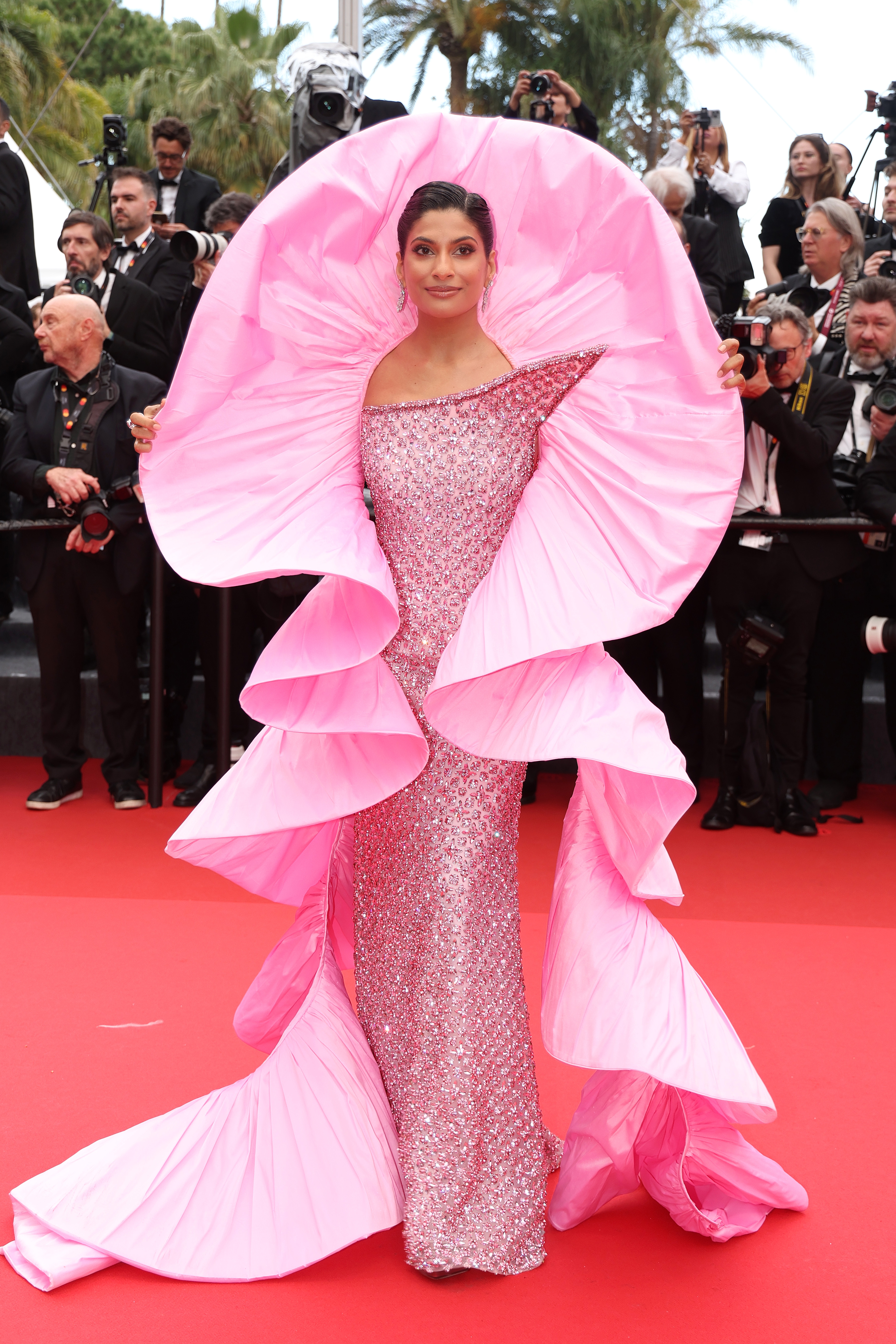 Farhana Bodi in an elaborate, layered gown with shimmer detailing, posing at a red carpet event, surrounded by photographers