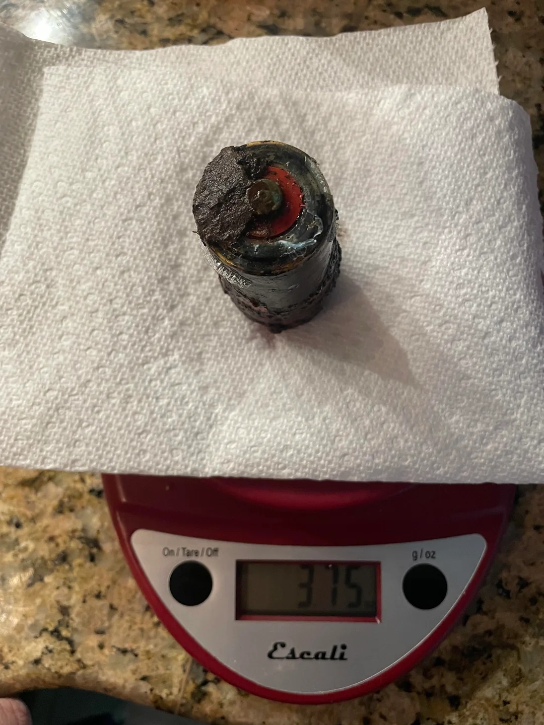A corroded battery on a digital scale reads 375 grams, with a paper towel underneath