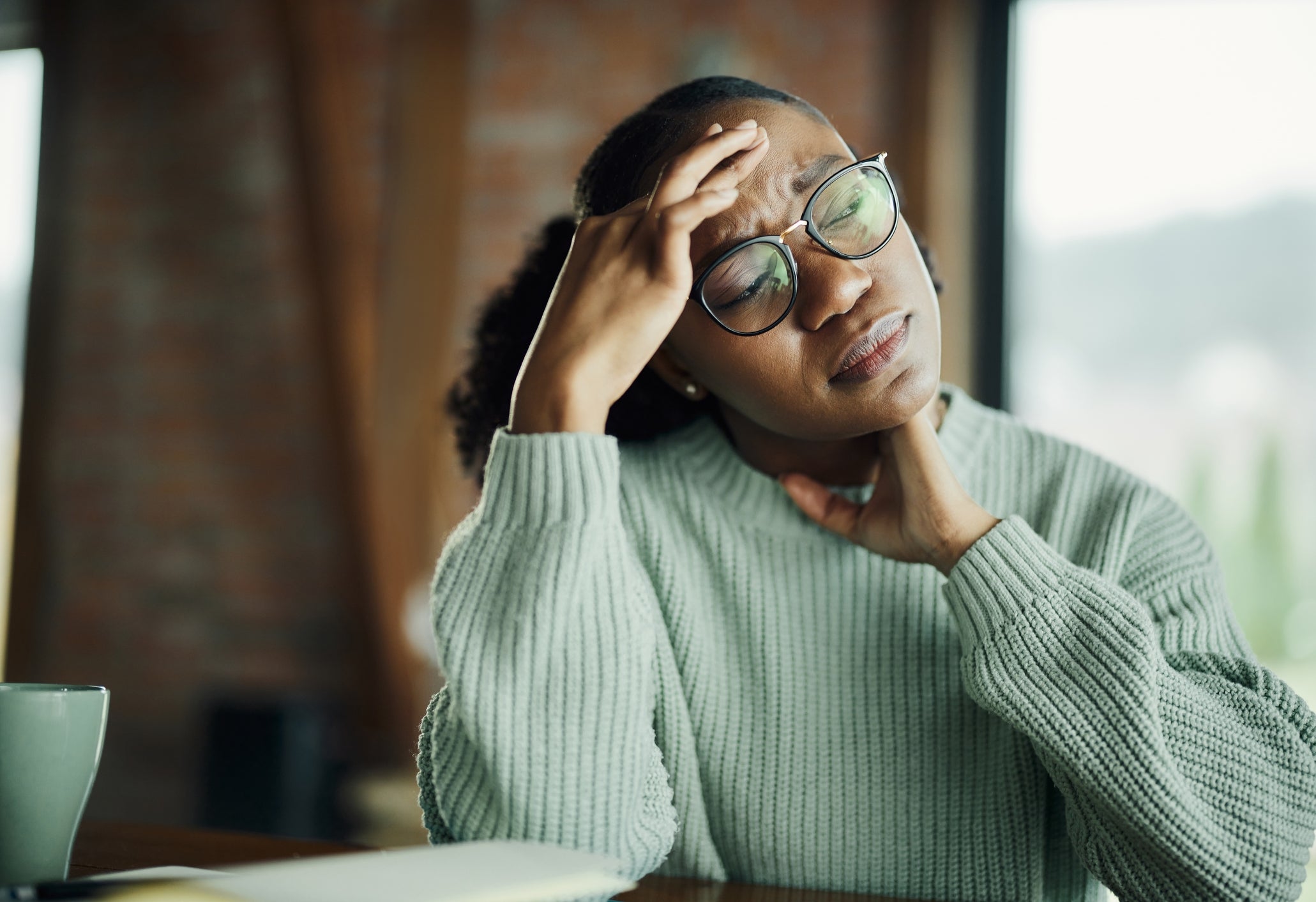 Person sitting at a table, touching their forehead with a pained expression, wearing glasses and a sweater