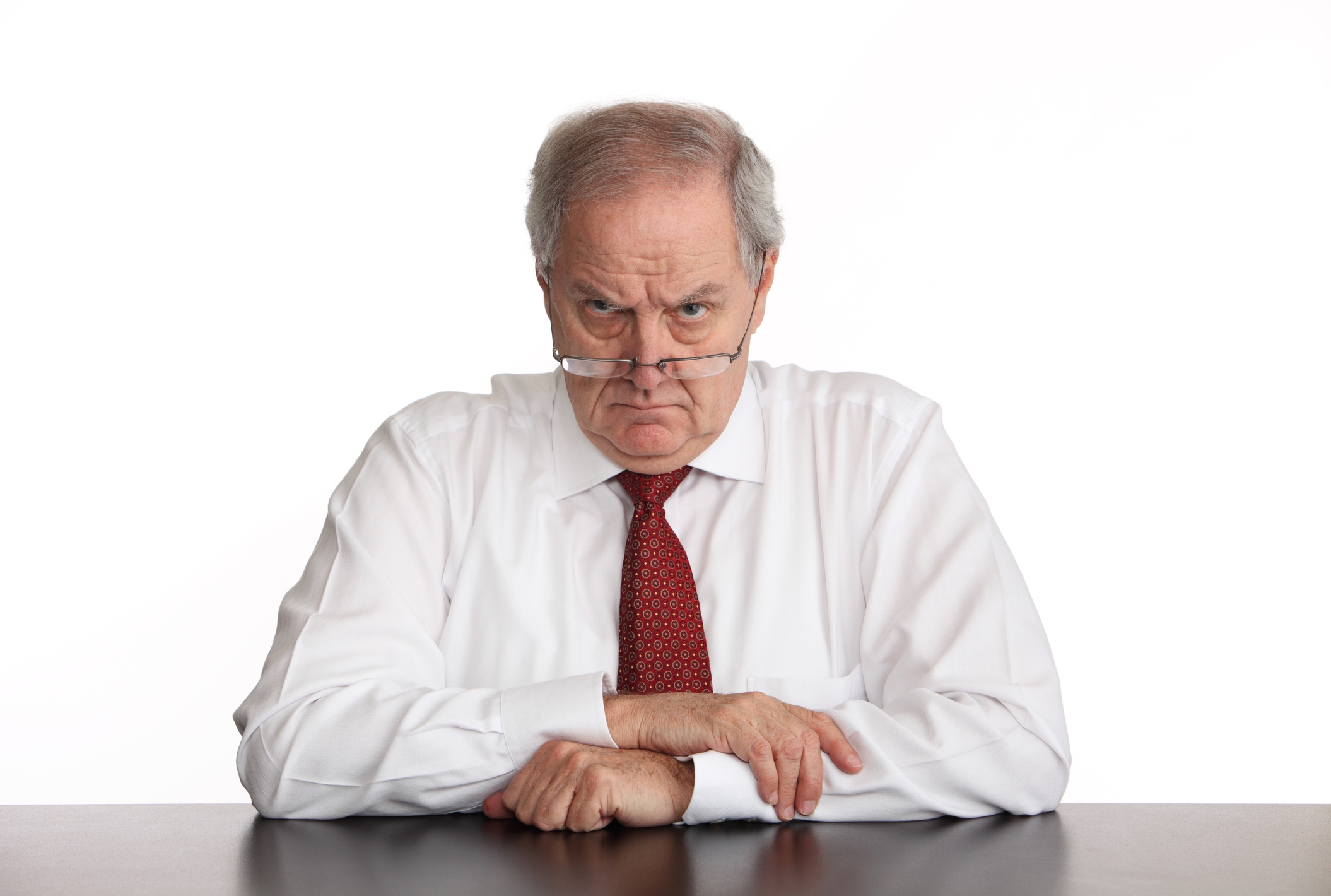Older man with glasses looks over them, wearing a white shirt and red tie, sitting at a table with a serious expression