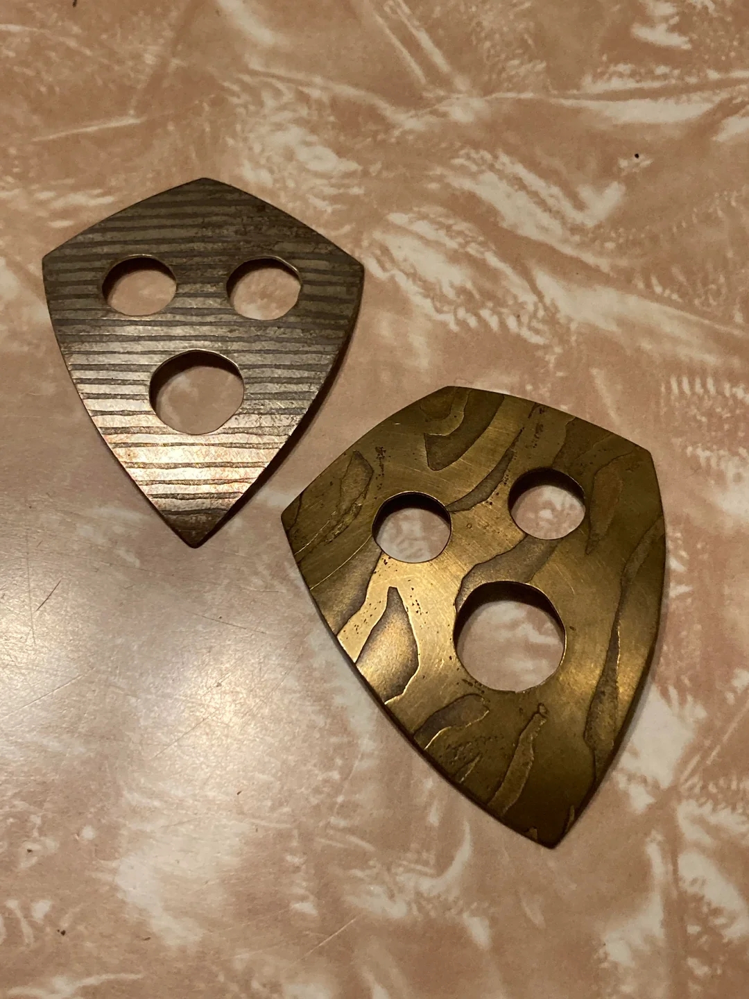 Two shield-shaped metal pieces with three circle cutouts each, placed on a textured surface