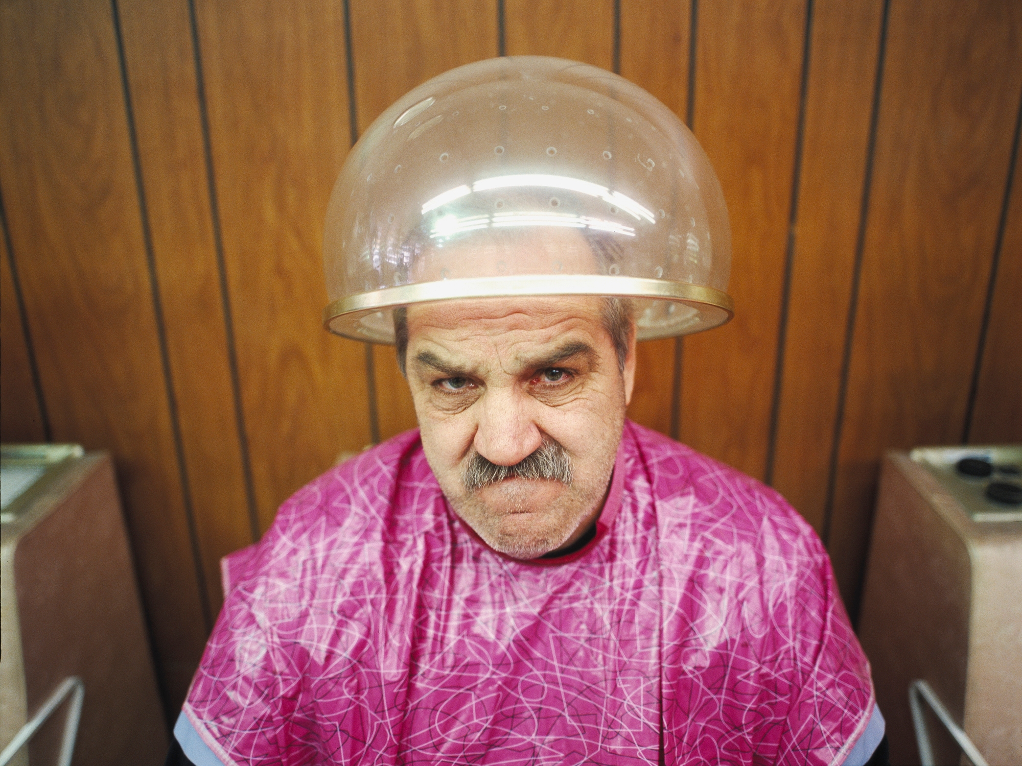 Man under hair dryer at salon, wearing a patterned smock, looking unimpressed